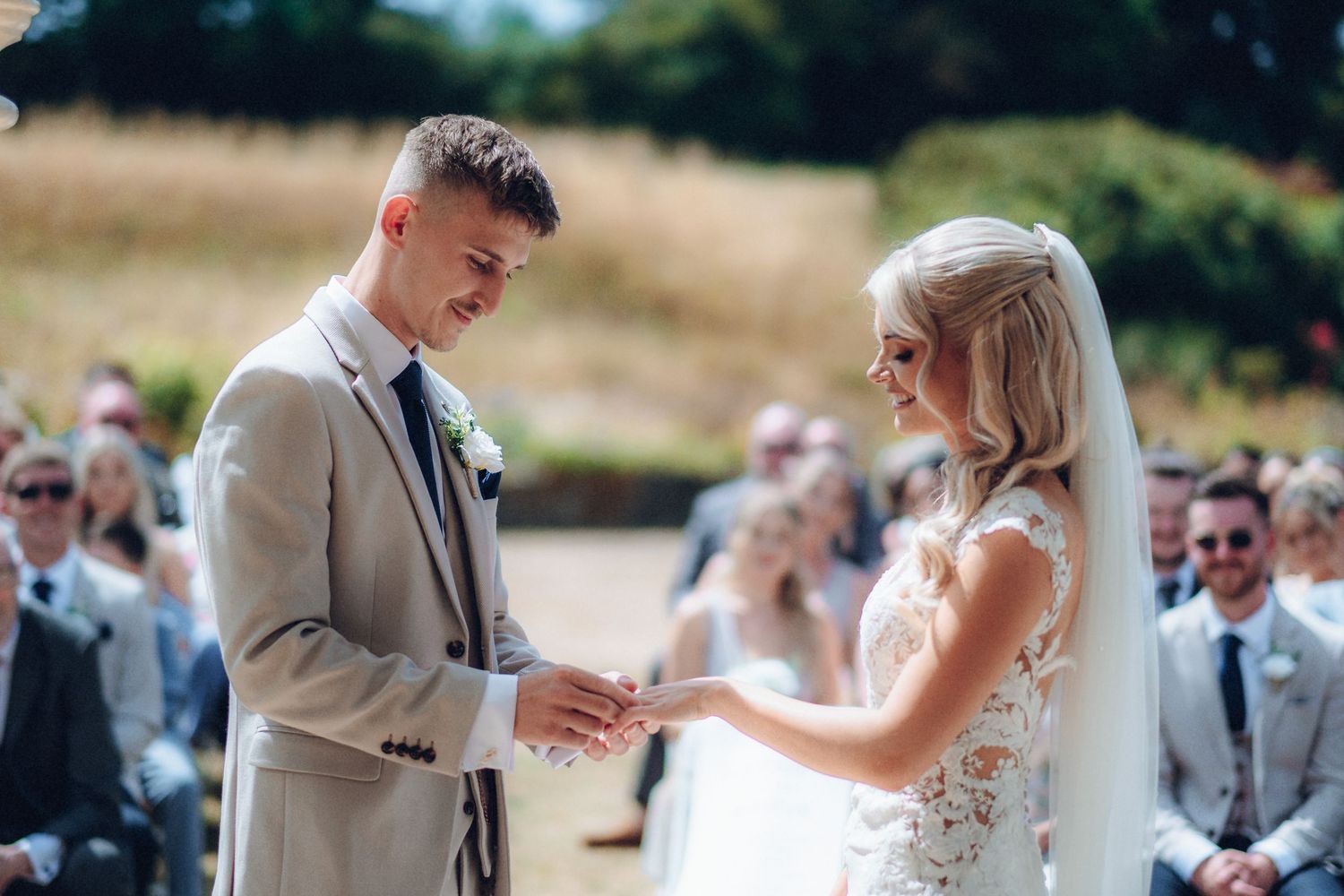 Couple exchanges rings during outdoor wedding ceremony with guests watching in background.