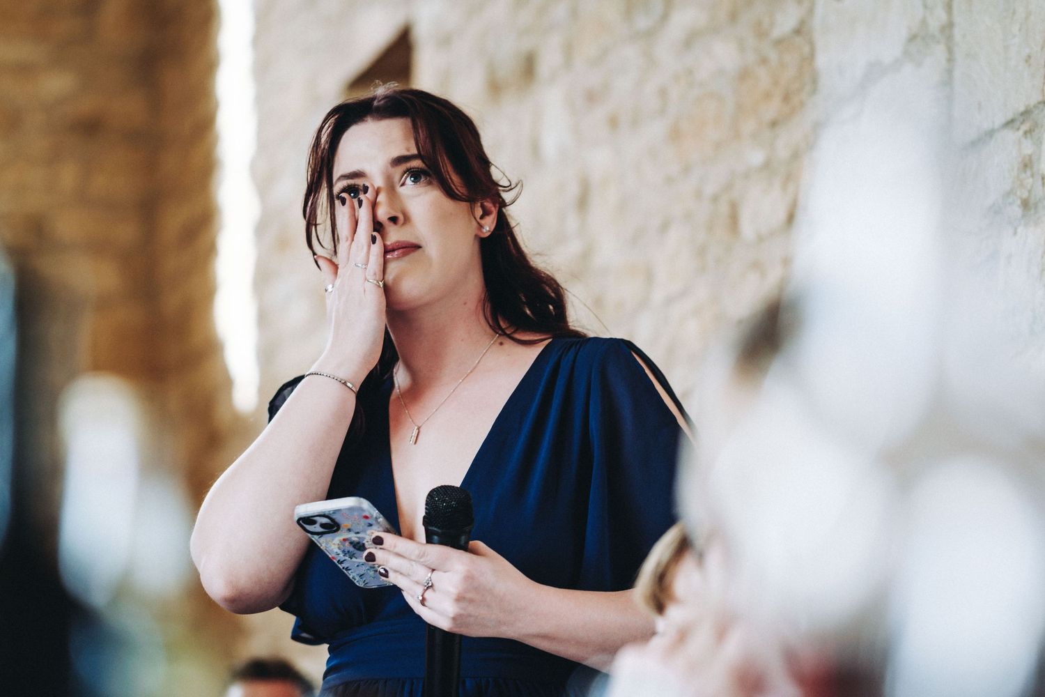 A person in a navy blue dress holds a camera while leaning against a stone wall in soft natural lighting.