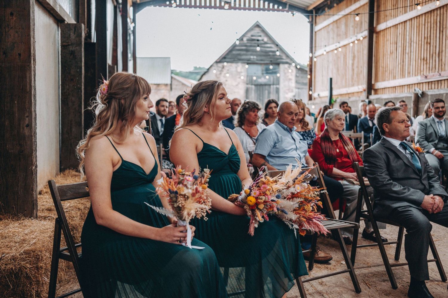 Wedding attendees in emerald green dresses holding autumn bouquets seated during outdoor Humber Barn ceremony.