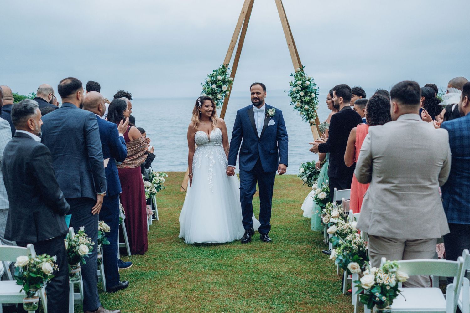 Wedding ceremony guests gathered around a wooden arch decorated with white flowers.