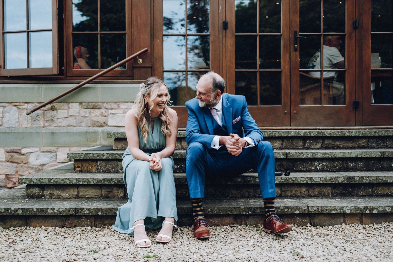 People gather on stone steps outside a building with large windows during a wedding event.