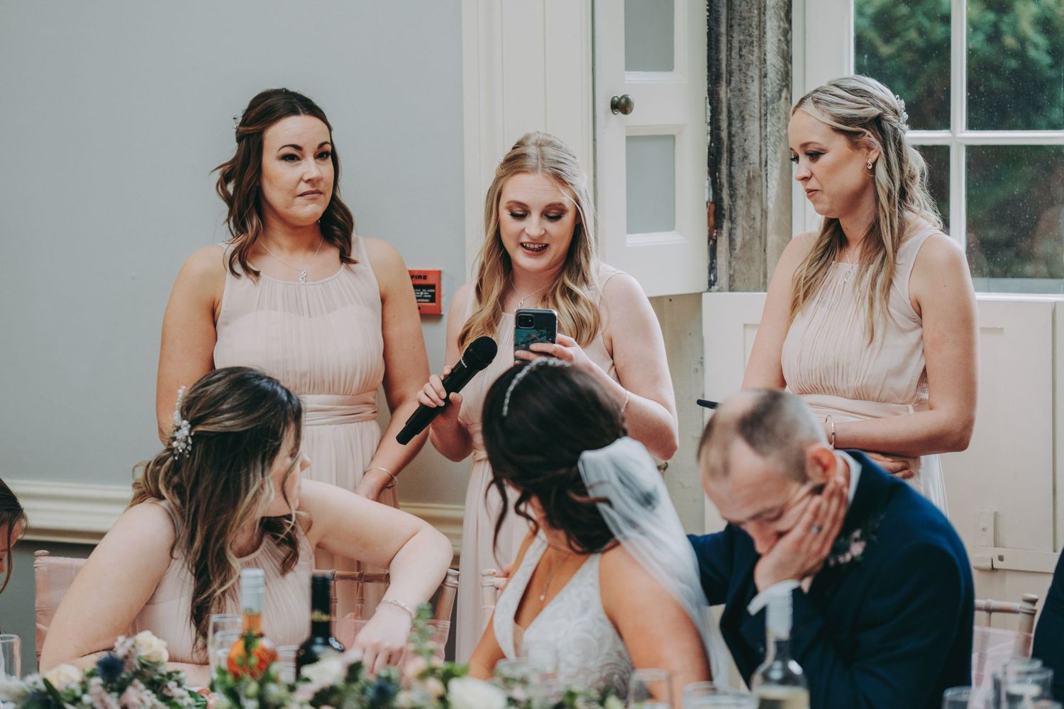 Bridesmaids in blush dresses gathered around table at wedding reception.