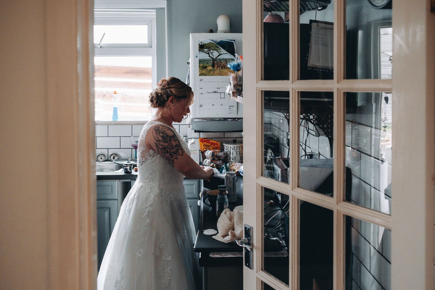 A figure in a white wedding dress stands in a doorway to a kitchen with white subway tile and shelving.
