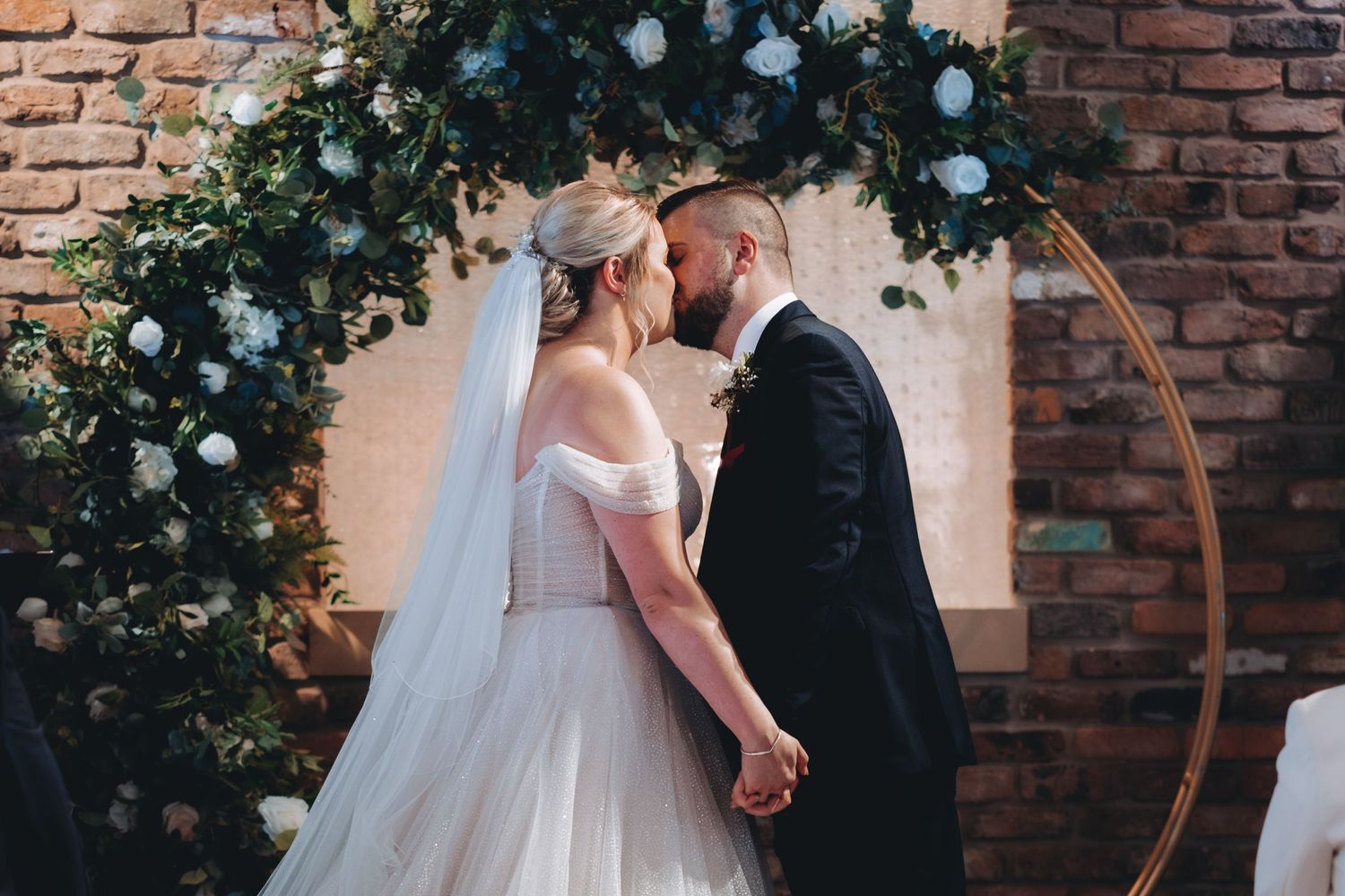Romantic wedding moment under a circular floral arch against rustic brick wall backdrop.