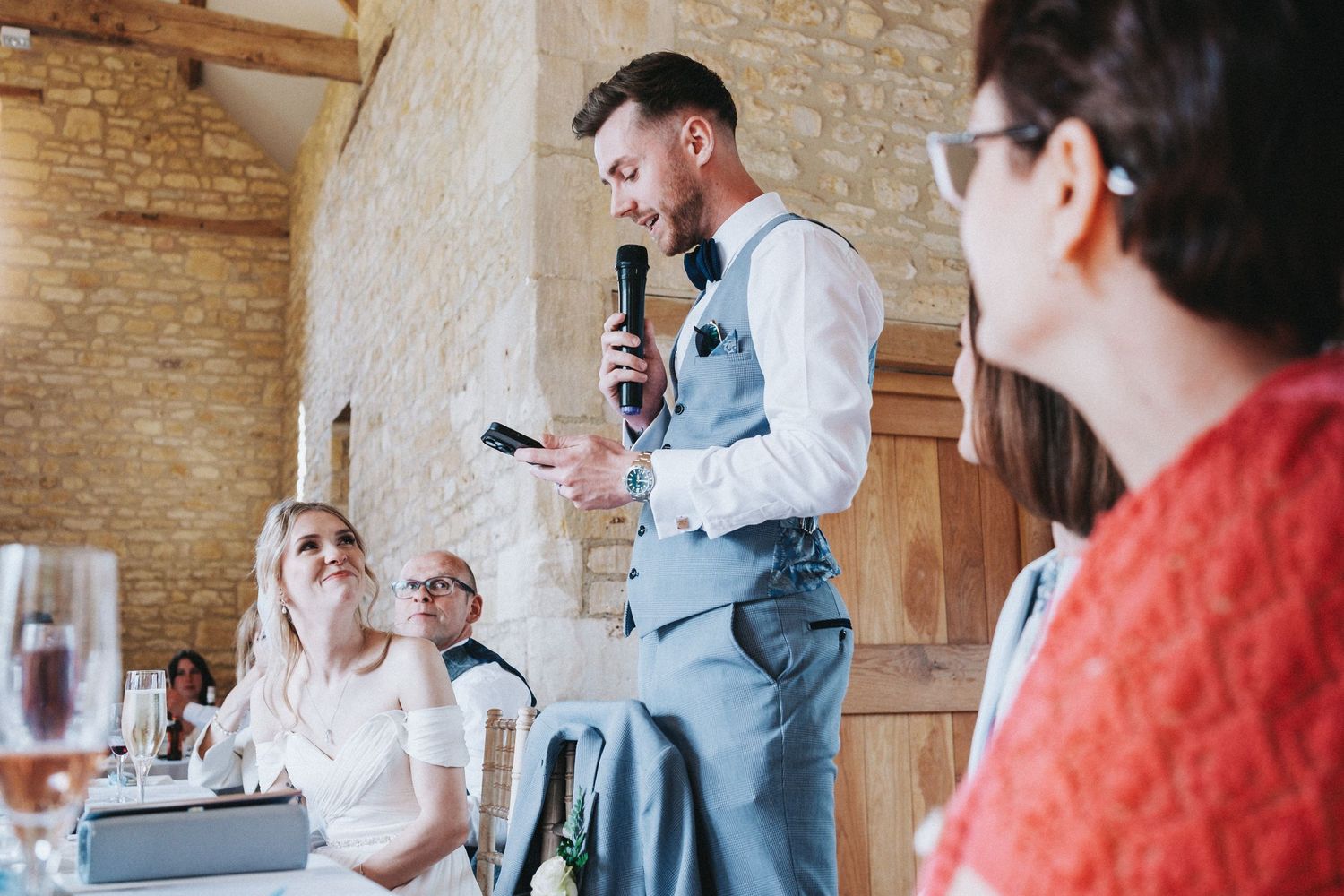 A wedding reception speech being delivered by a guest wearing a vest and bowtie in a rustic stone venue.