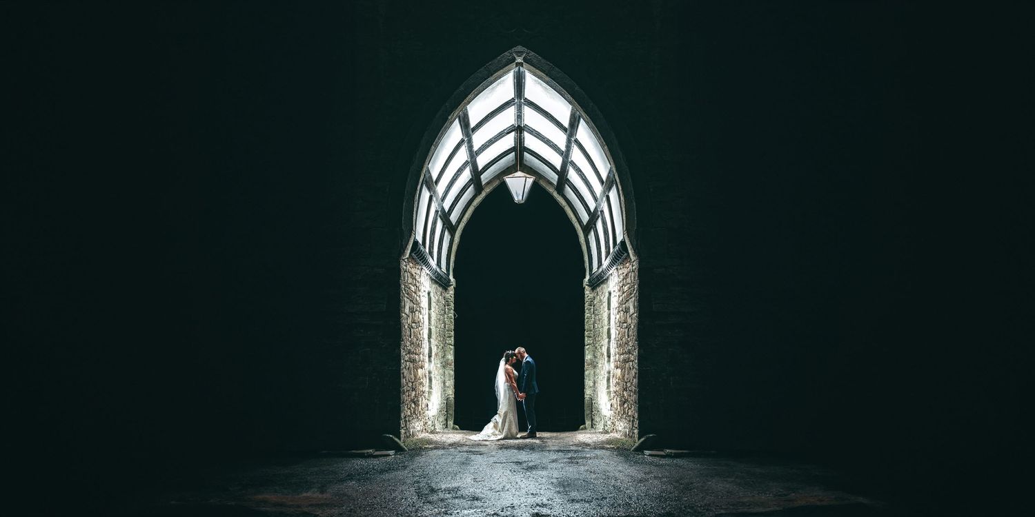 A dramatic wedding photo captured through an illuminated gothic archway against a dark background.
