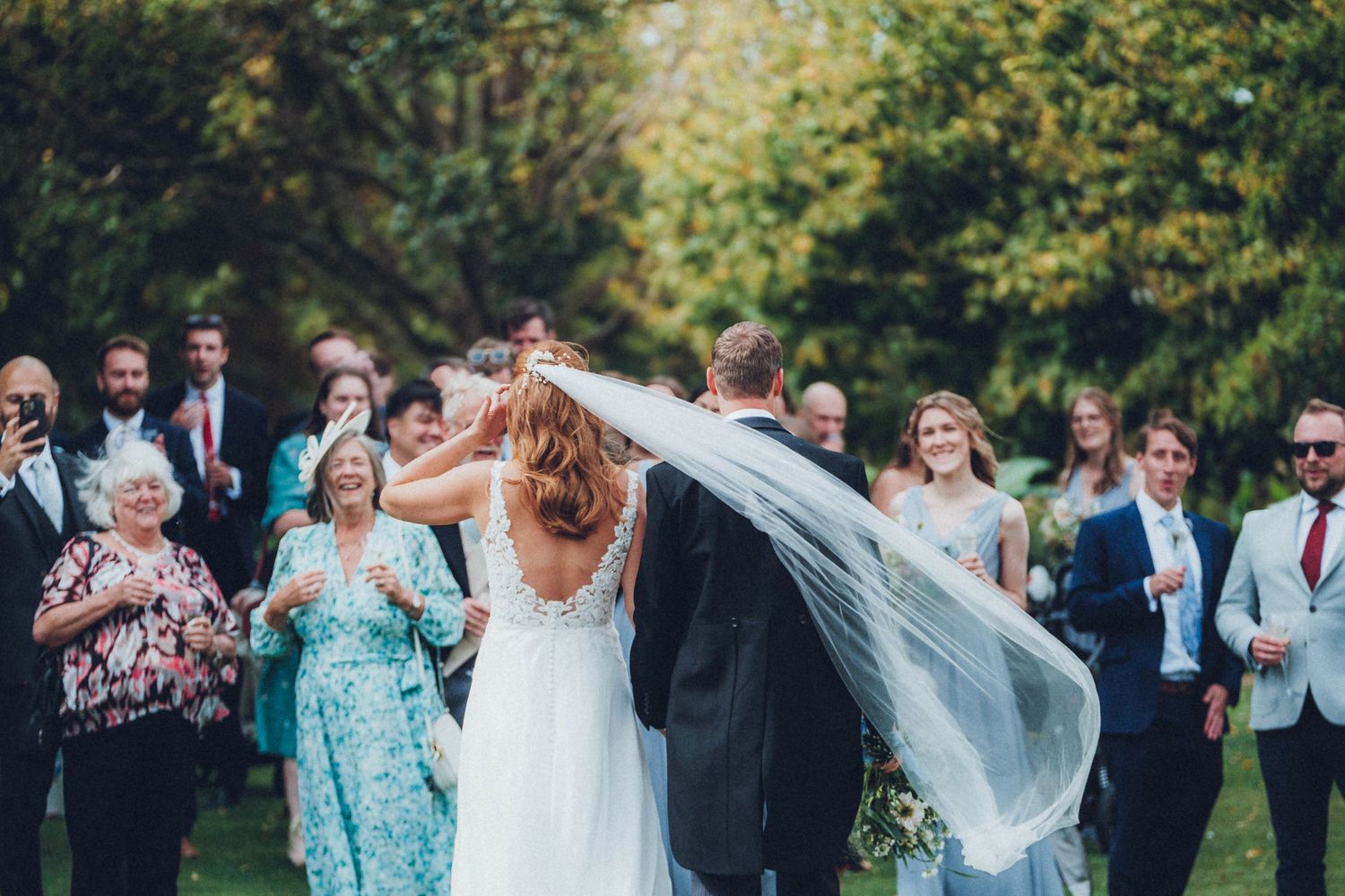 Outdoor wedding celebration with flowing veil and guests cheering in natural setting.