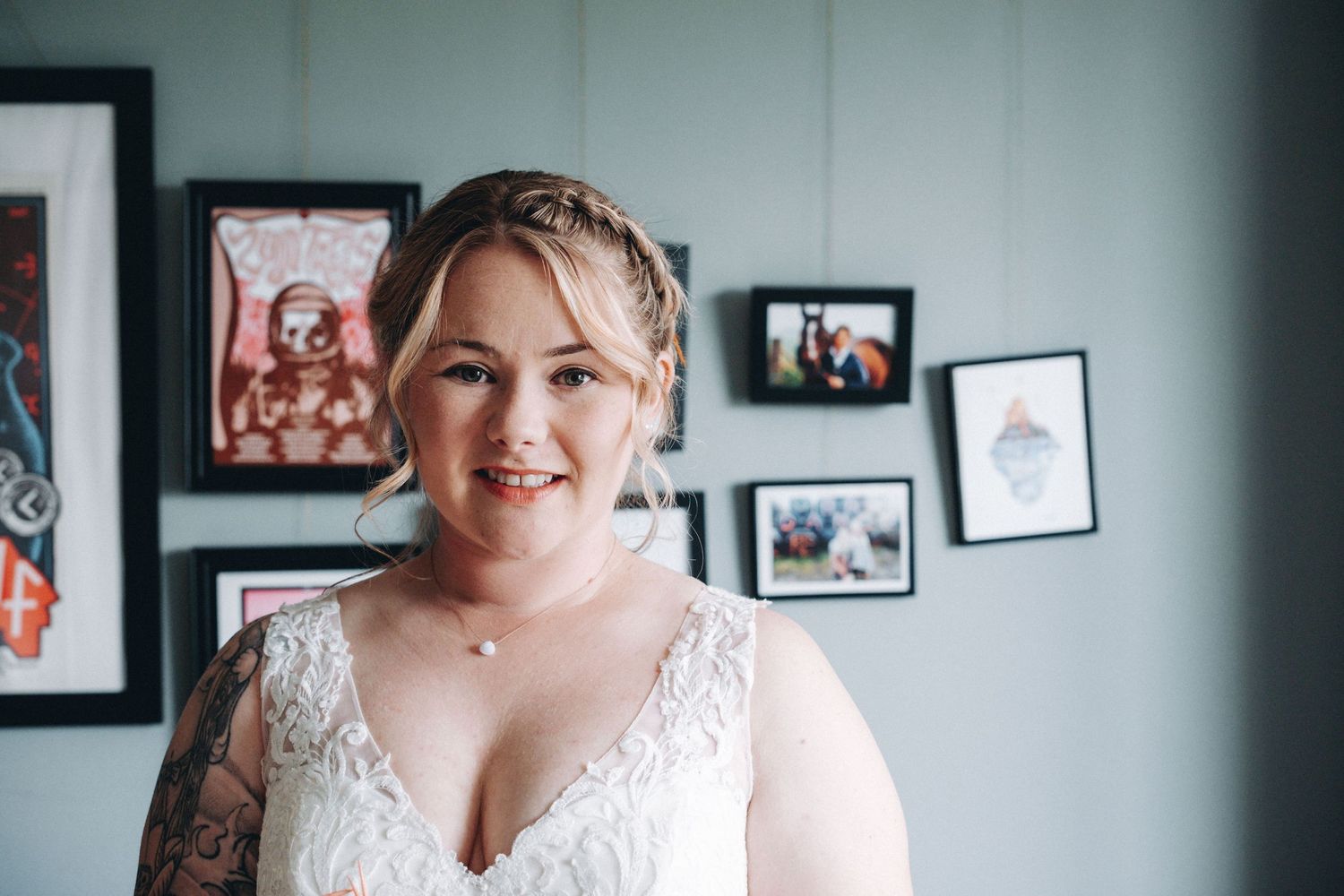 A bride in a white lace wedding dress smiles while standing in front of framed photos on a gray wall.