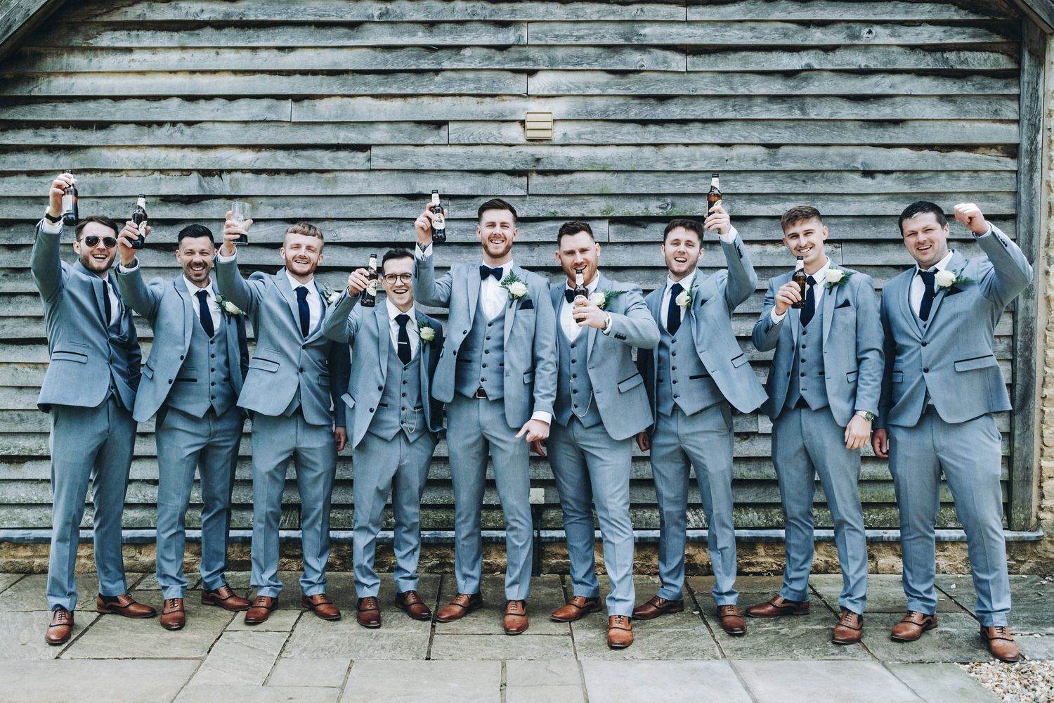 Groomsmen in matching light gray suits and brown shoes pose casually against rustic wall background.