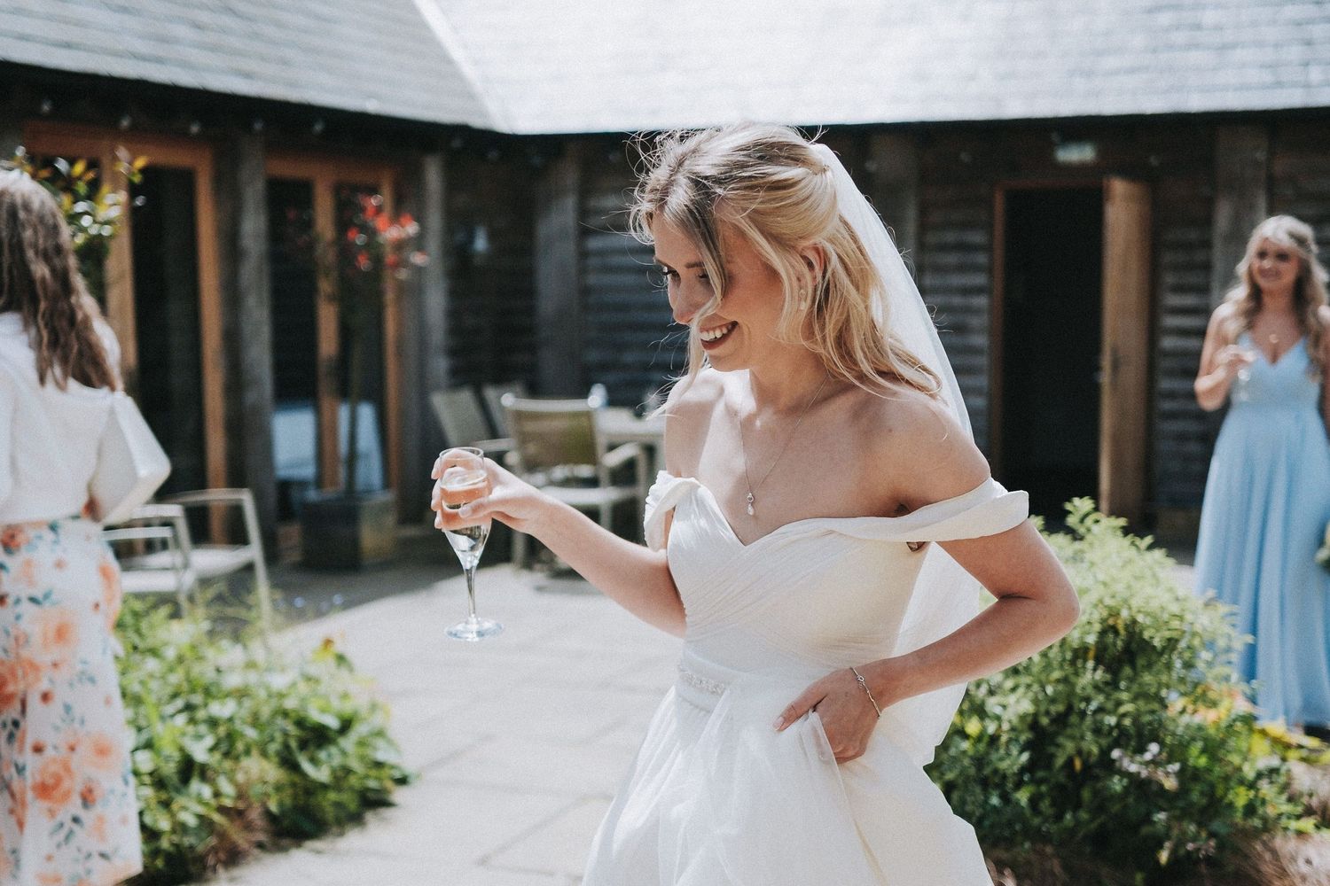 A bride in an off-shoulder white gown holds a champagne glass in an outdoor garden setting.
