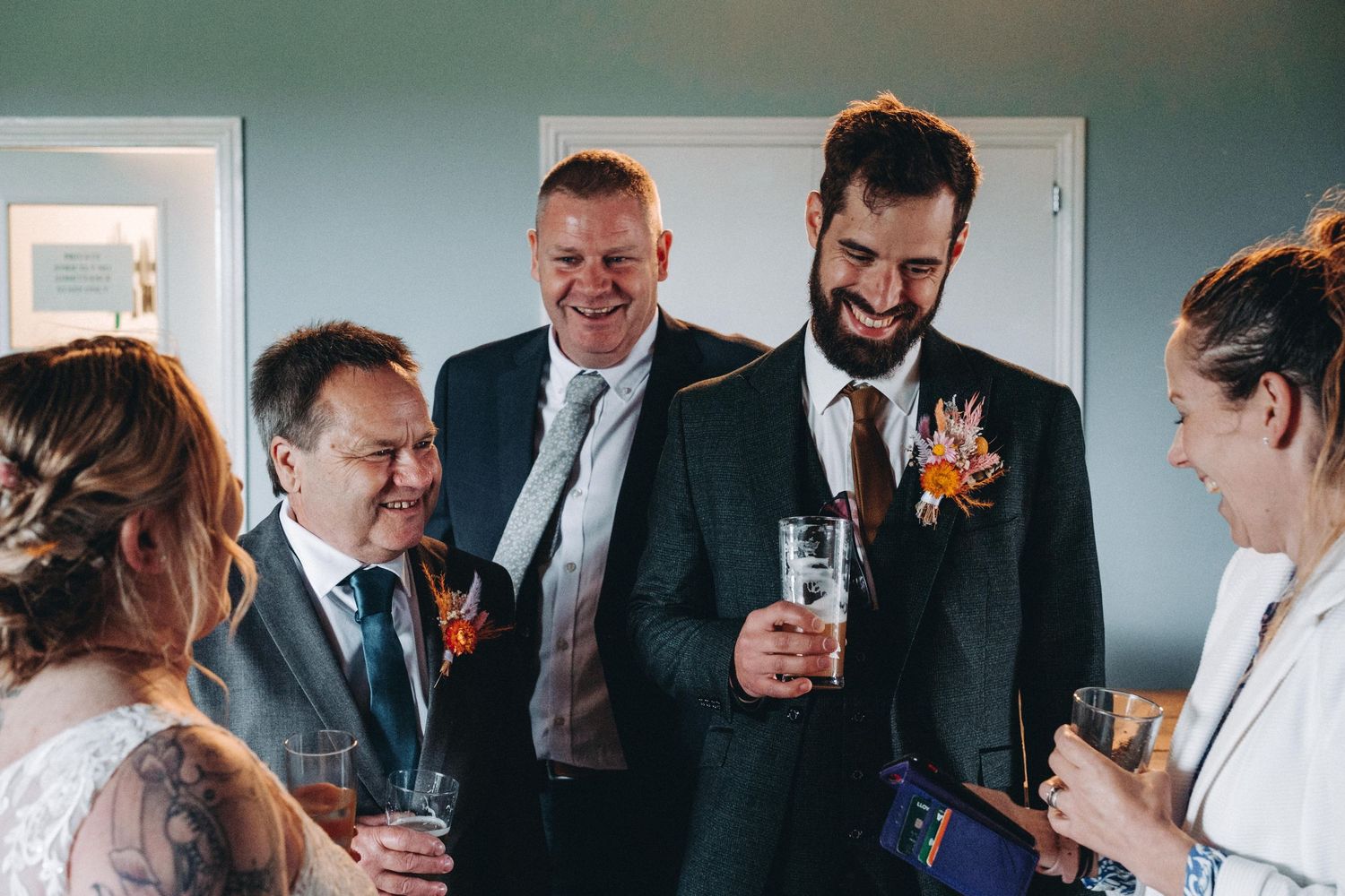 Wedding party members in dark suits with orange boutonnieres share drinks and laughter during a celebratory moment.