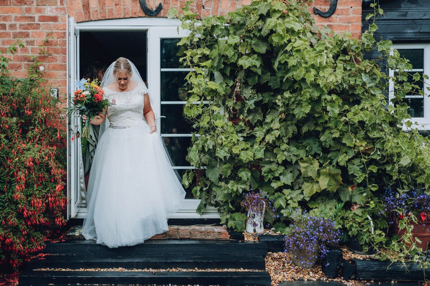 Bride in white wedding gown standing on steps with lush garden foliage.