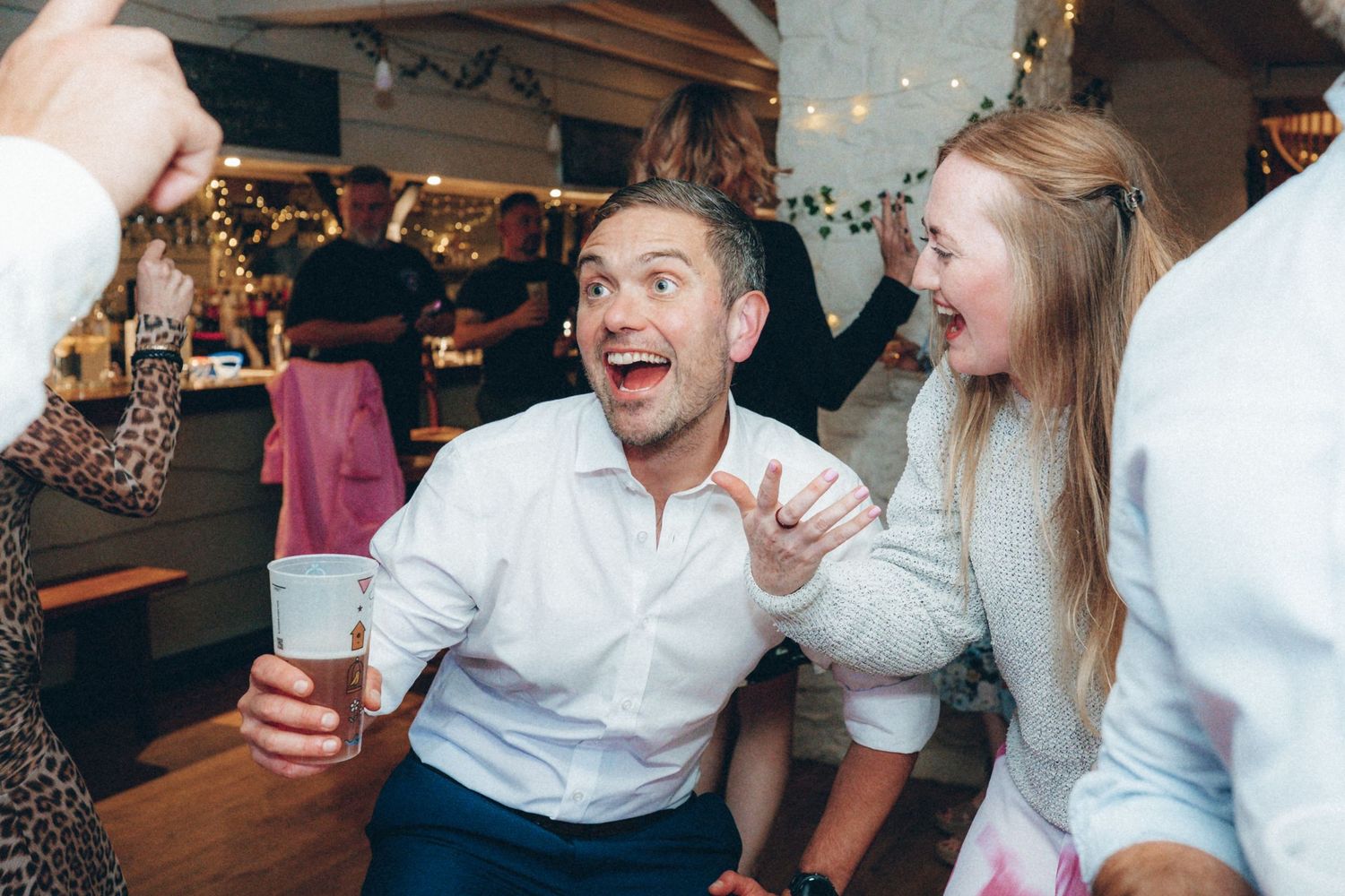 Party guests dancing and laughing during a celebration in a rustic white-walled venue.