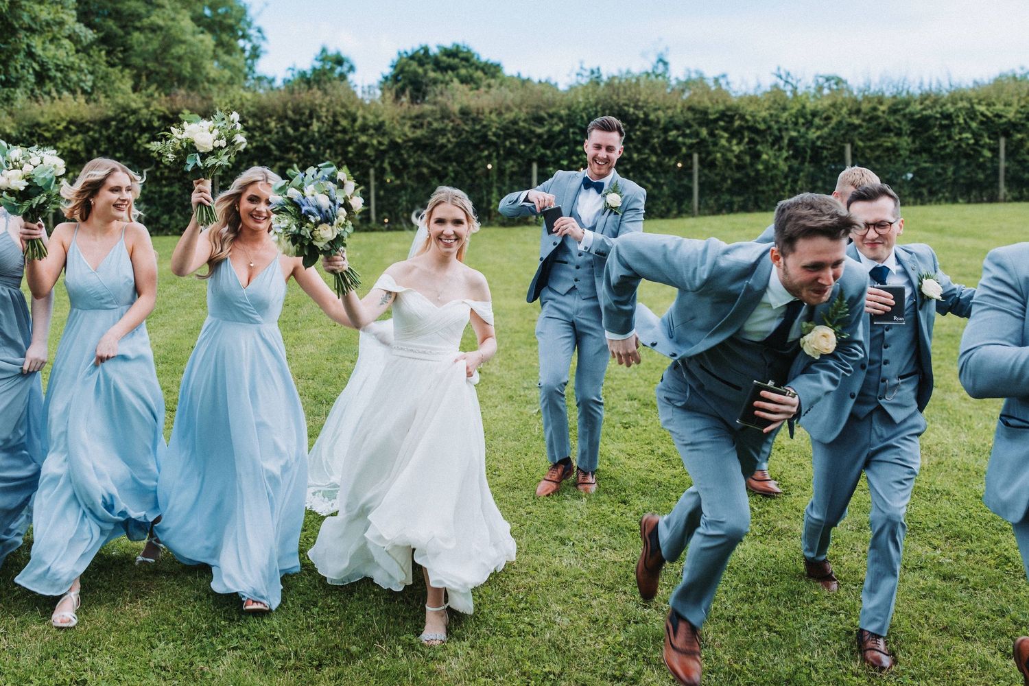 Wedding party members in light blue bridesmaid dresses and gray suits run playfully across a grassy lawn during celebration.