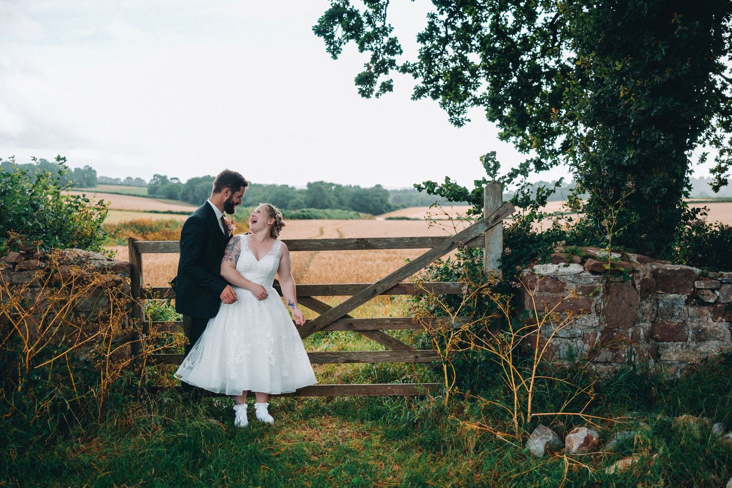 A couple in formal wedding attire poses by a rustic wooden gate in the countryside with fields stretching behind them.