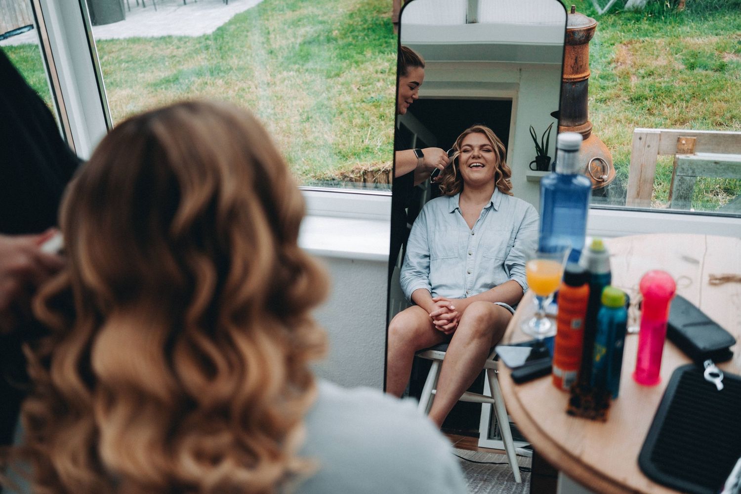 A bridesmaid getting hair styled in a makeup chair with beauty products visible in a mirror reflection.