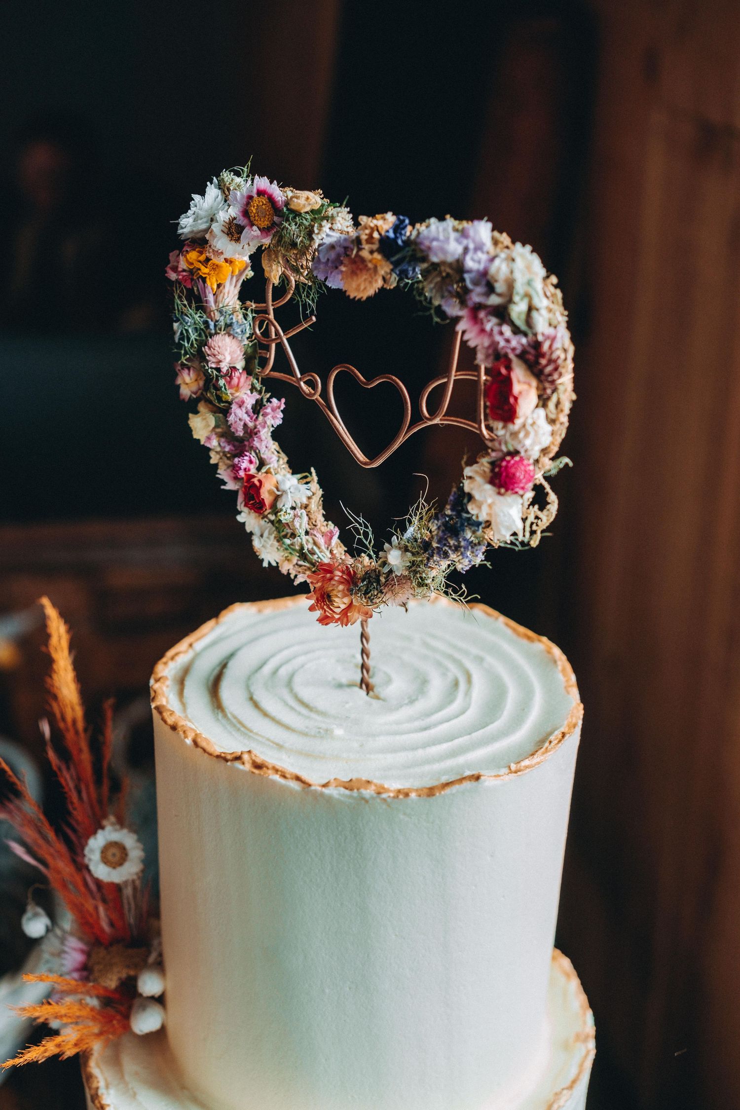 White wedding cake topped with heart-shaped dried flower wreath cake topper featuring delicate pink and purple blooms.