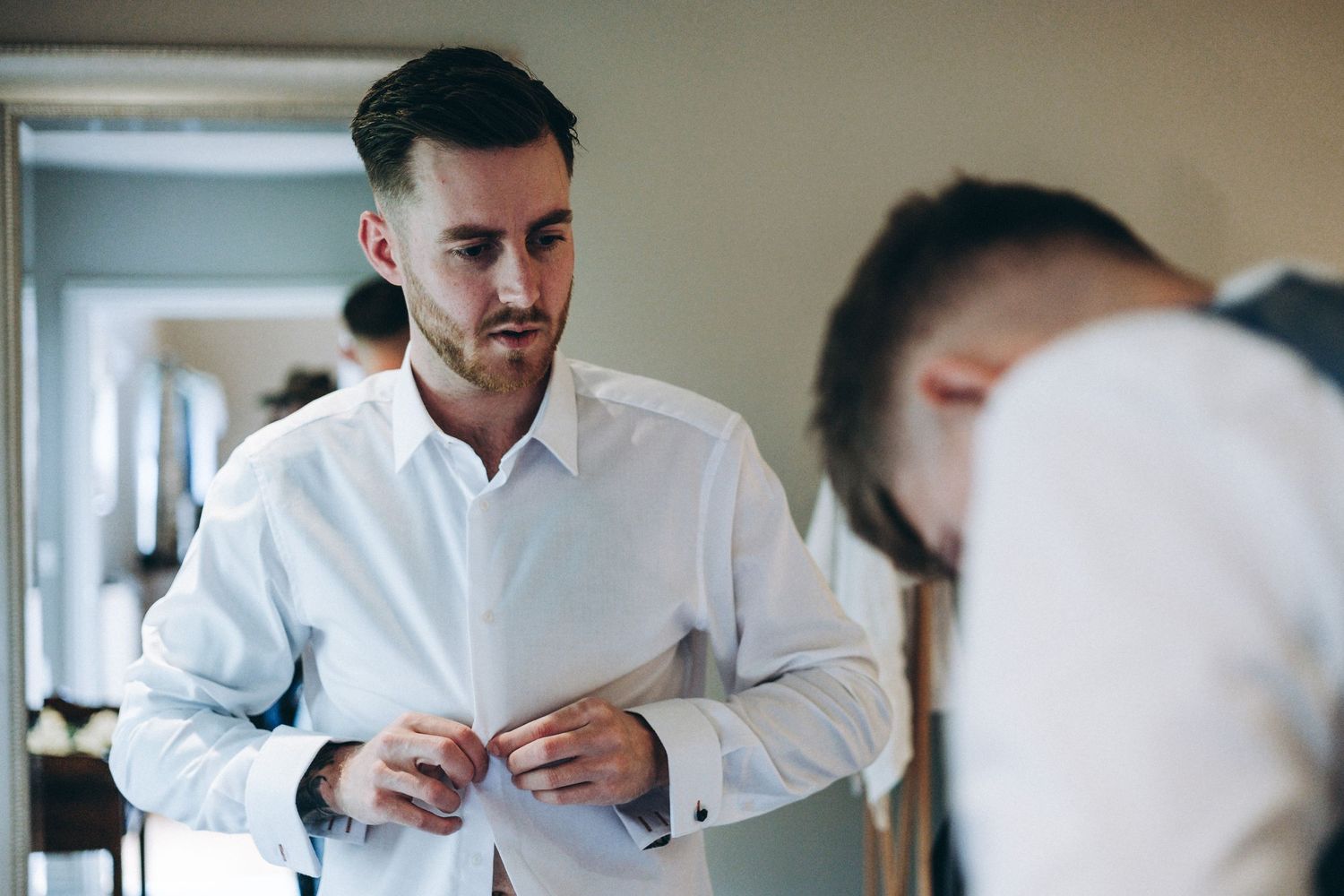 Groom in a white button-down shirt adjusts his clothing while getting ready in a hallway with soft lighting.