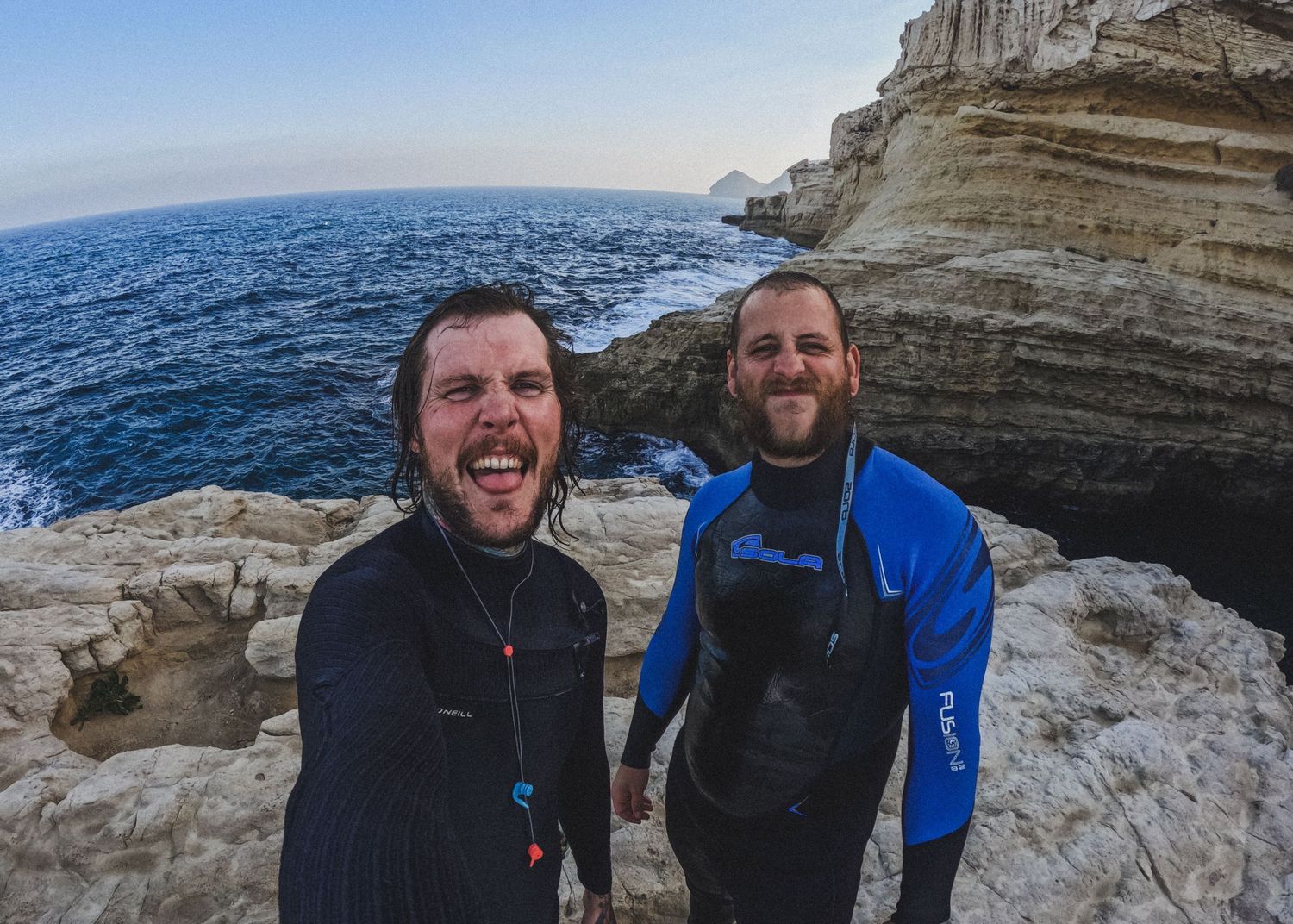 Two people smiling at camera while standing on rocky ocean cliffs.