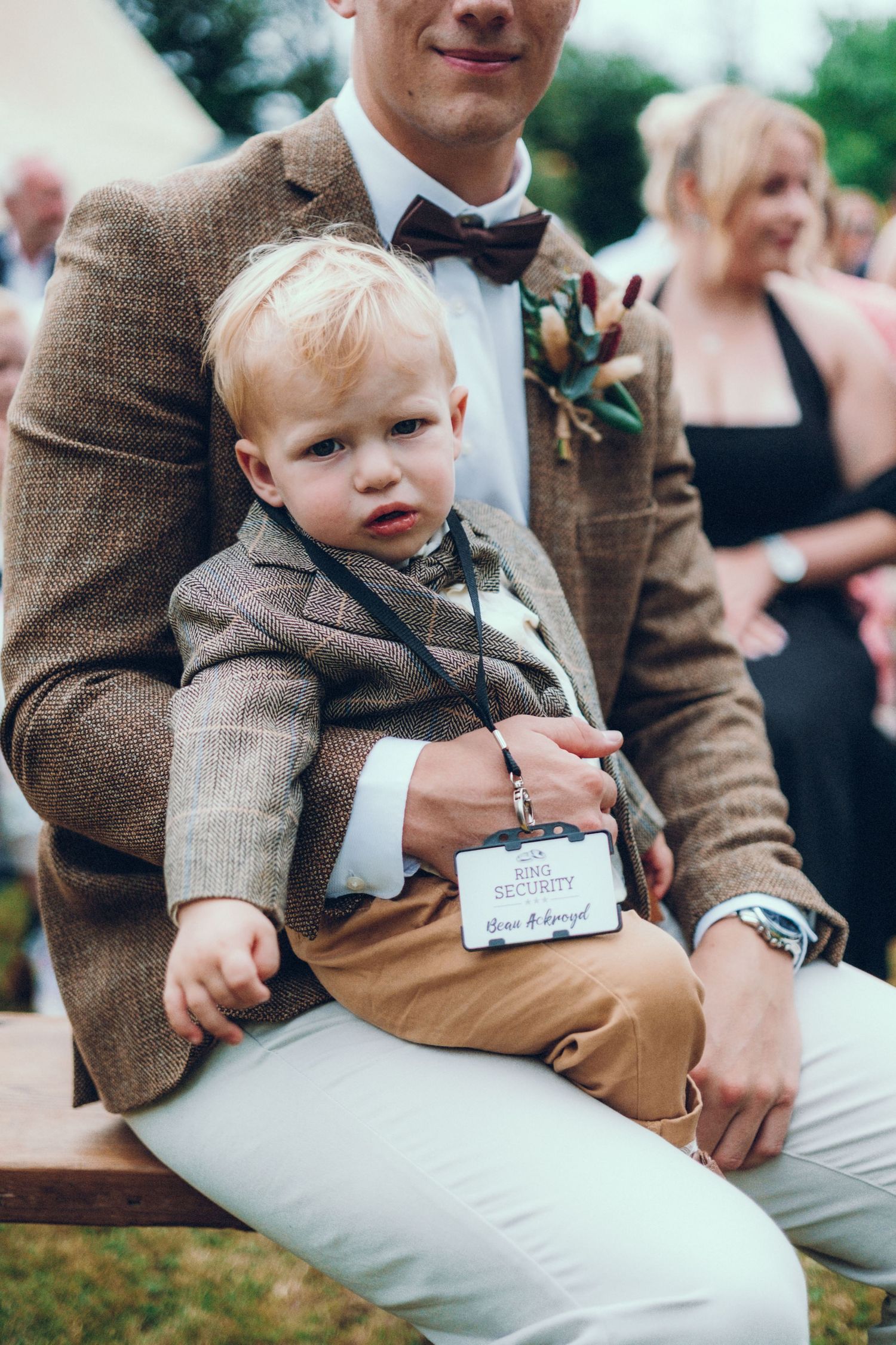 Young child in formal brown jacket sitting on adult's lap at outdoor event.
