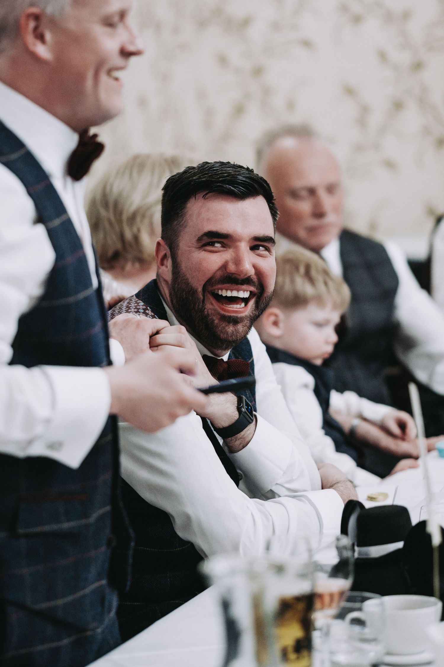 Wedding reception guests laugh and celebrate at dining tables.