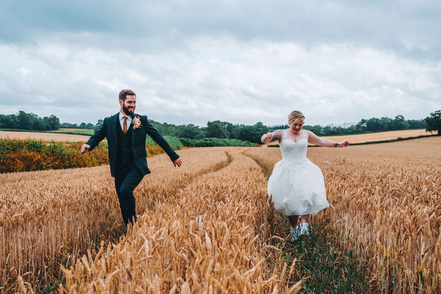Newly wed couple runs joyfully through a golden wheat field on their wedding day against a cloudy sky. Humber Barn, Devon Wedding.