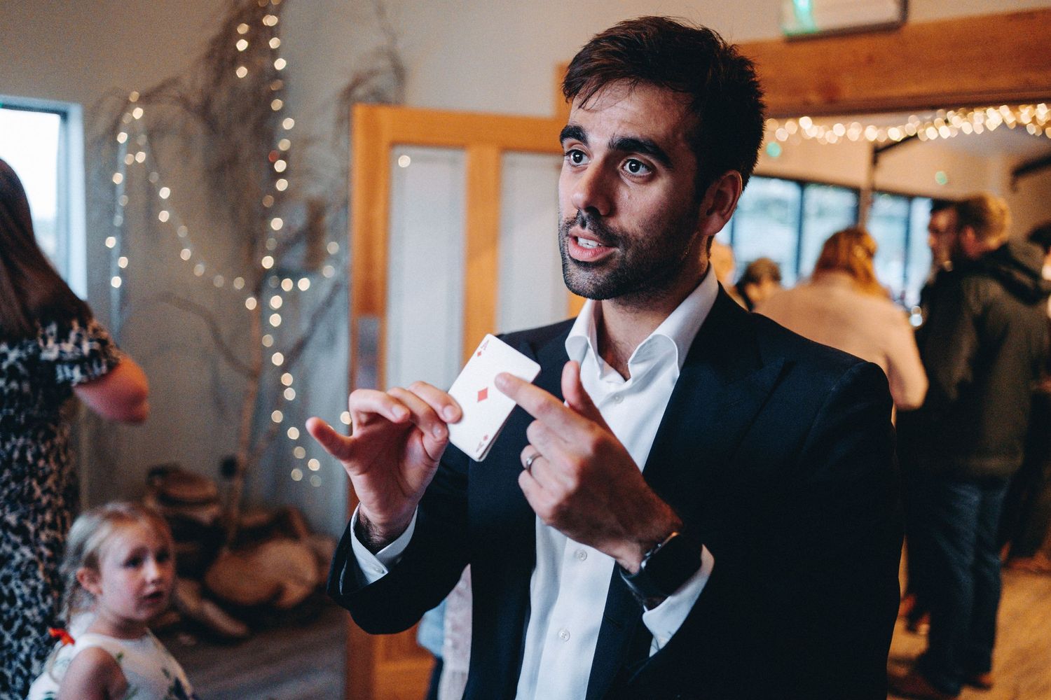magician in a black suit gestures while speaking at an event with string lights and decorations in the background.