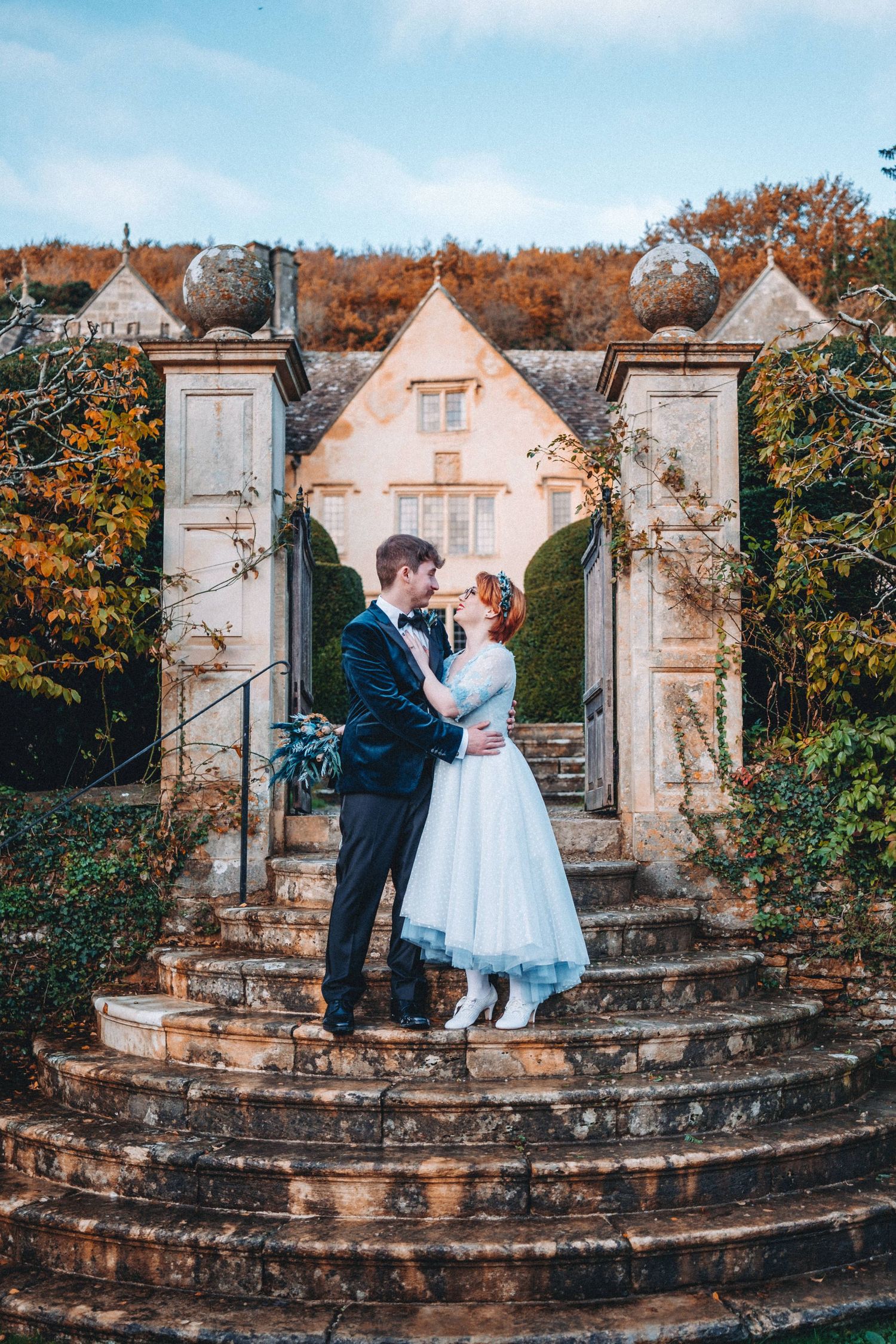Romantic wedding portrait on stone steps with ornate pillars and autumn foliage.