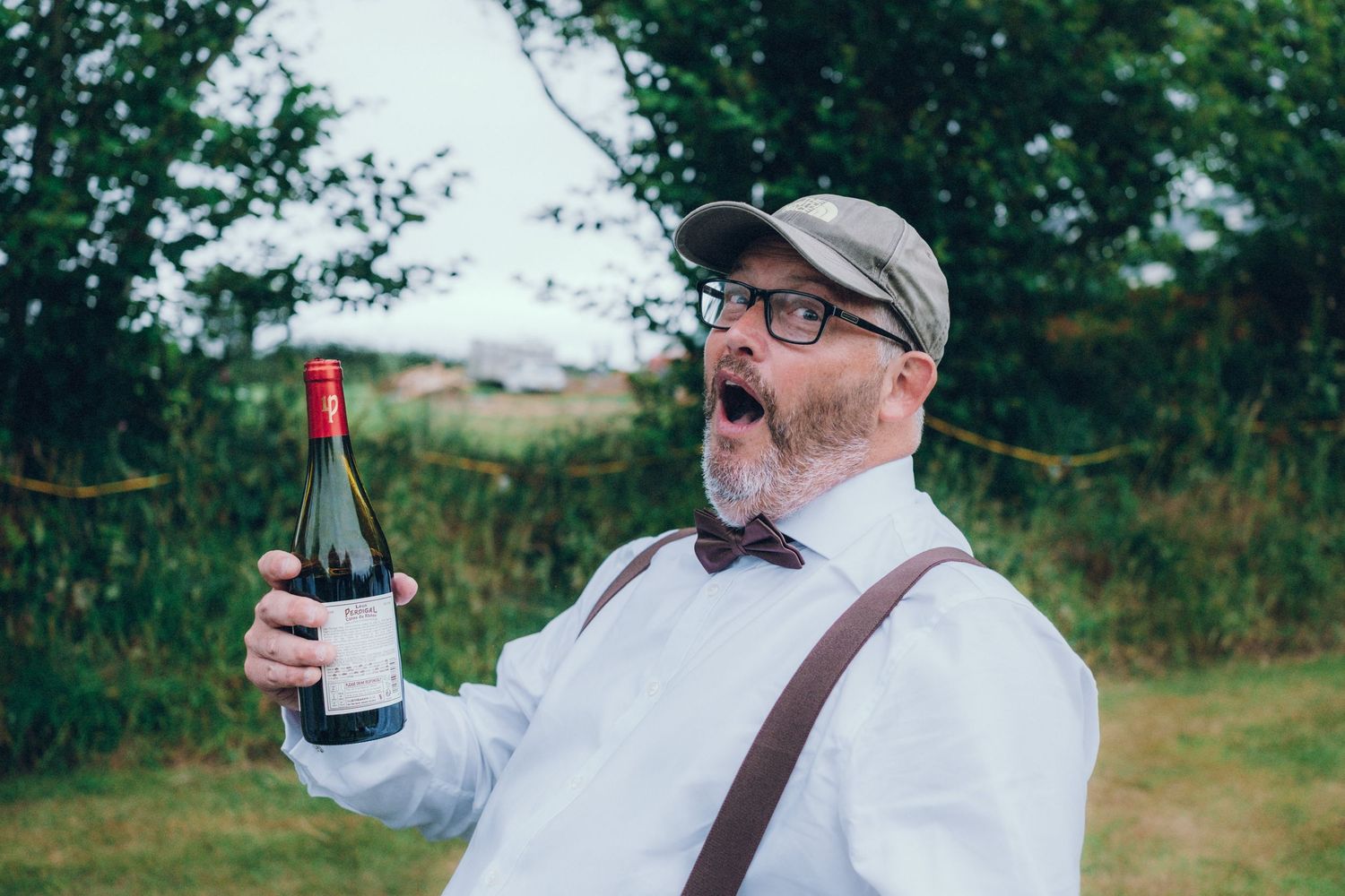 Guests in suspenders and bowties enjoy drinks at an outdoor wedding reception.