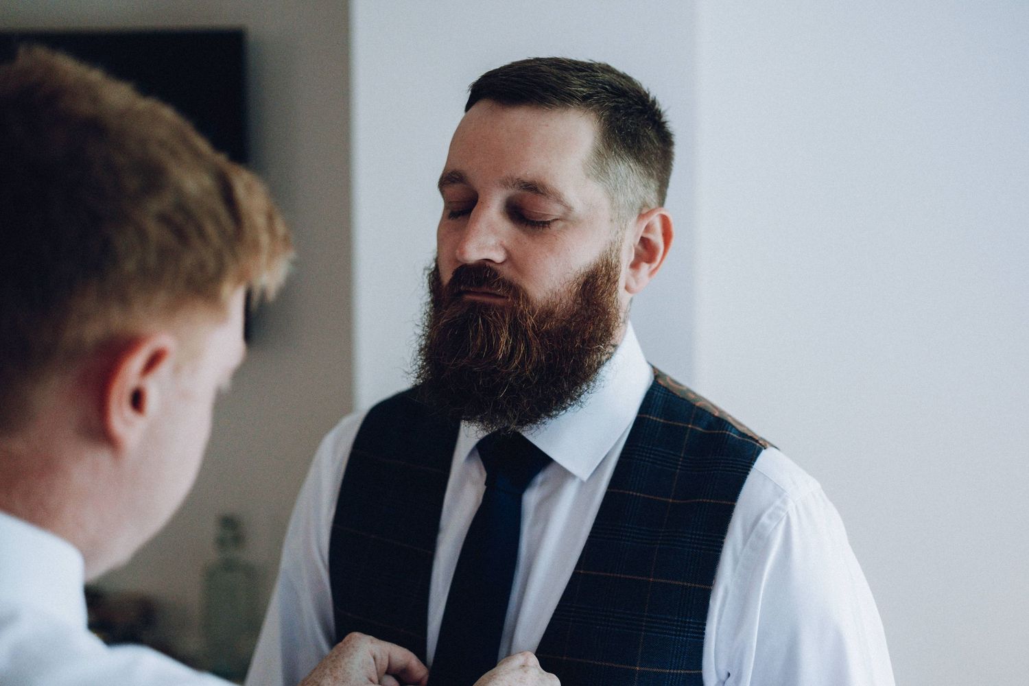 A bearded man in a suit and tie adjusts his cuffs while getting ready in a bright room.