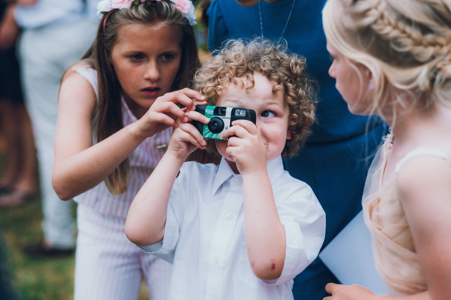 A young child at a wedding taking photos with a small film camera while others look on.