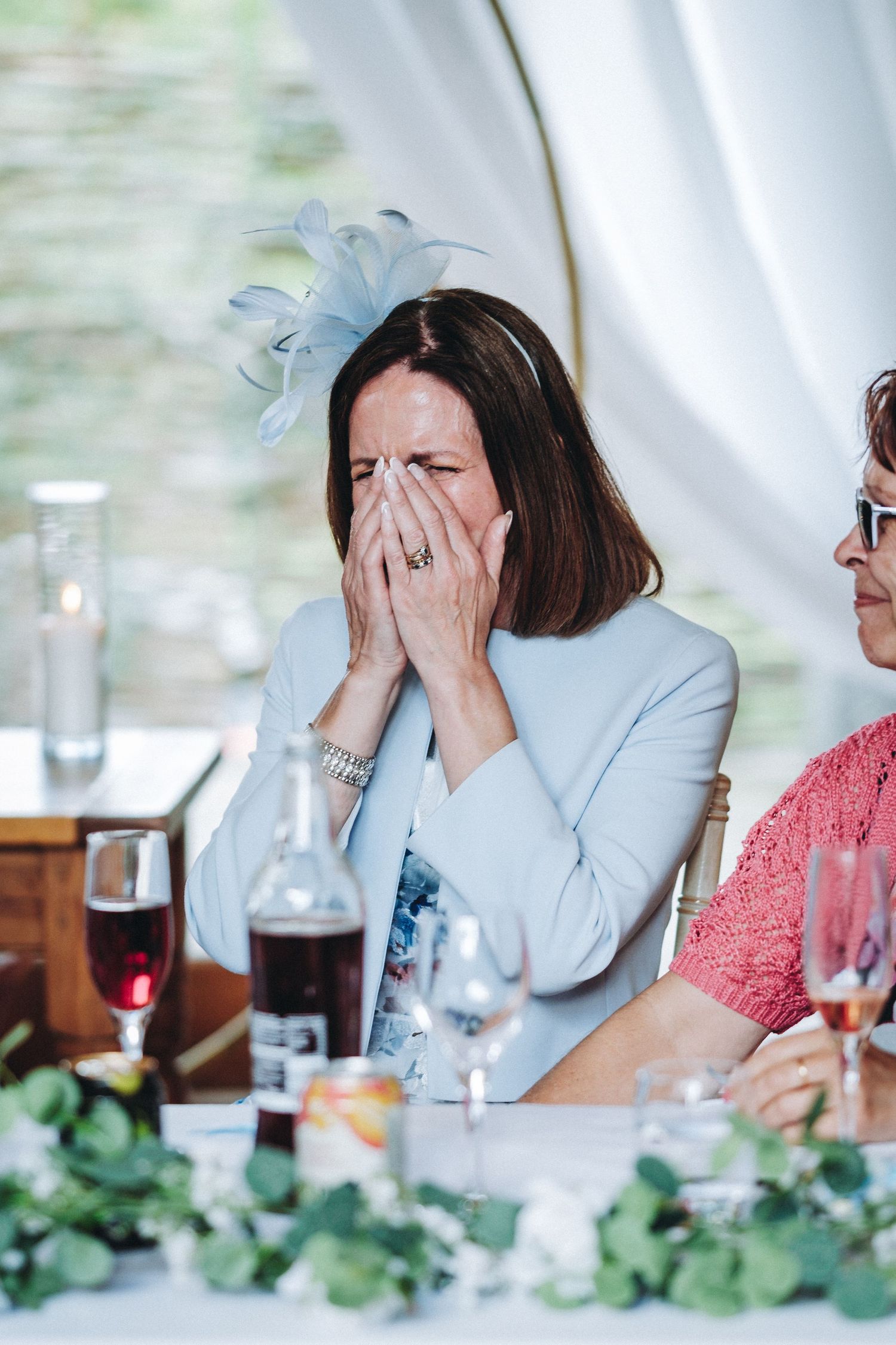 Wedding guest in light blue dress laughs with hands covering face at table decorated with greenery.