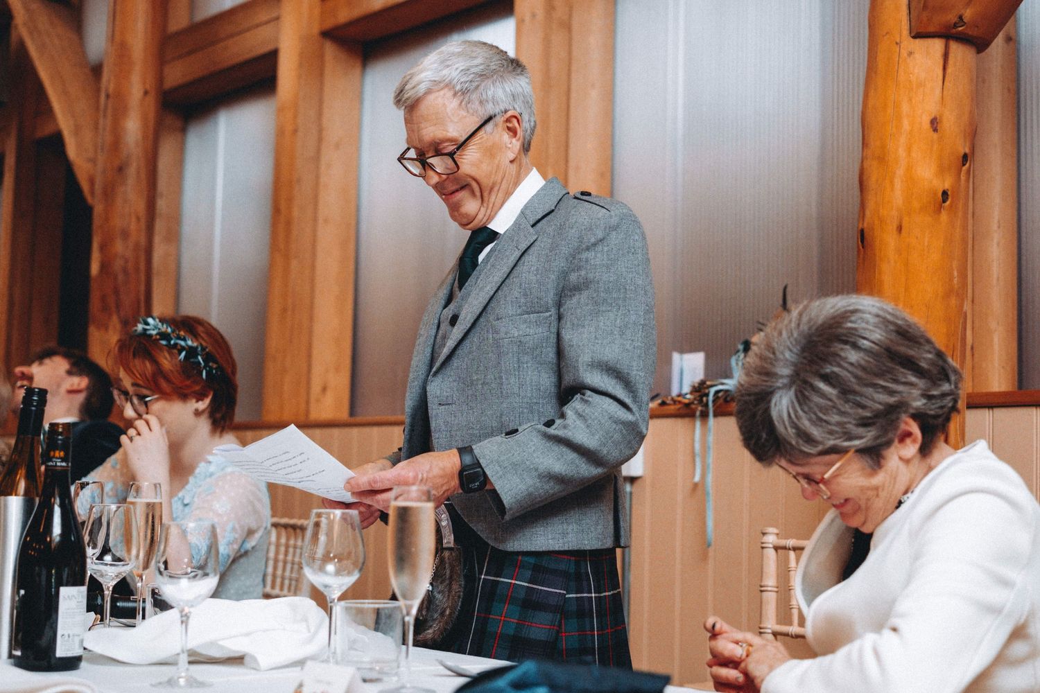 Wedding reception dinner scene with guests seated at tables in a wooden-beamed venue.