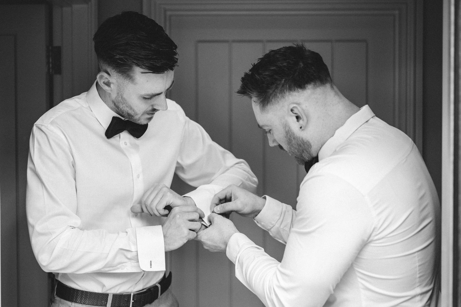 A black and white photo of two people in formal attire helping each other with cufflinks before a special event.