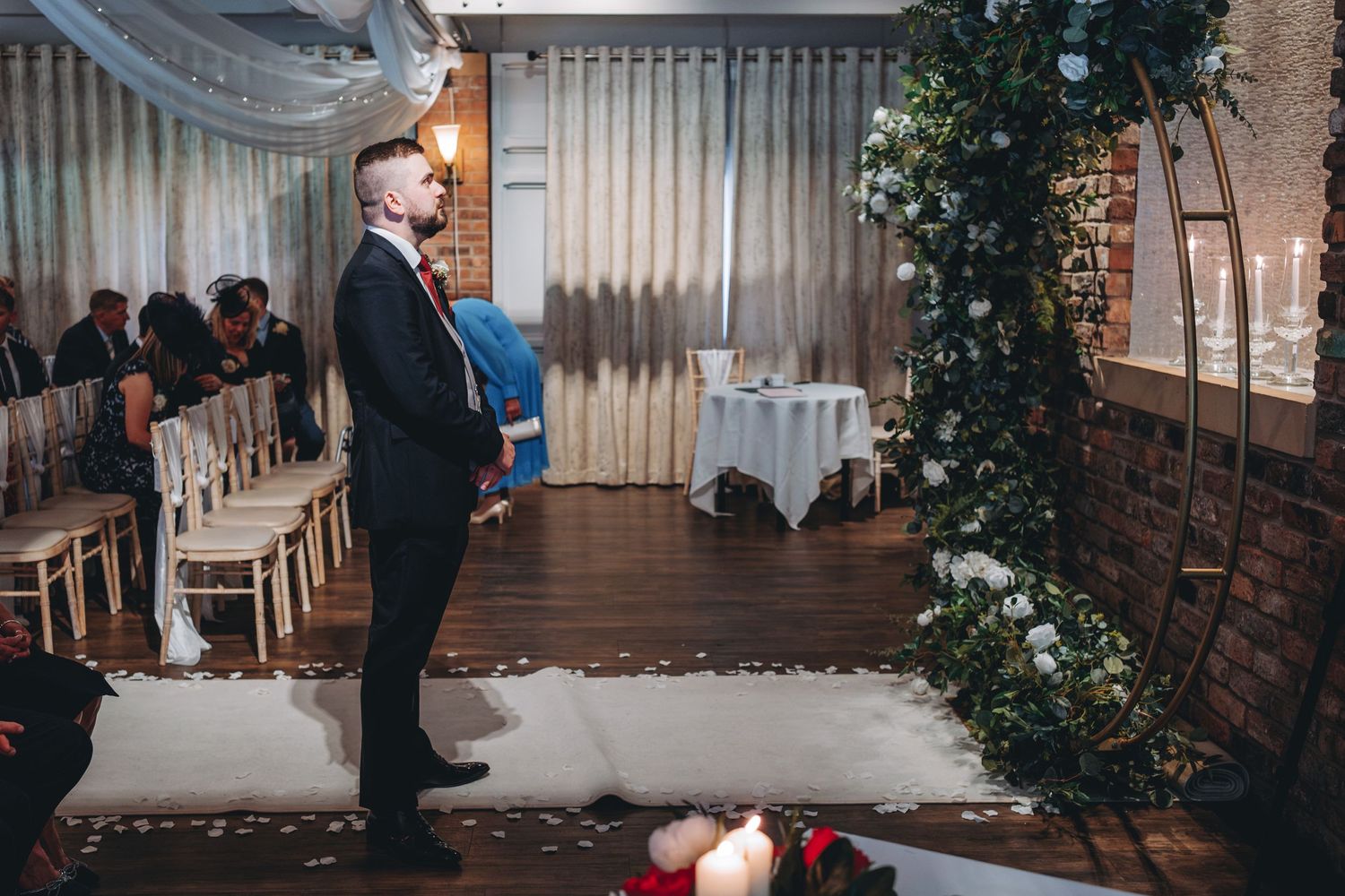 Groom waits patiently.
Elegant wedding venue with festive decorations featuring brick walls, white chairs, and floral arrangements at sunset