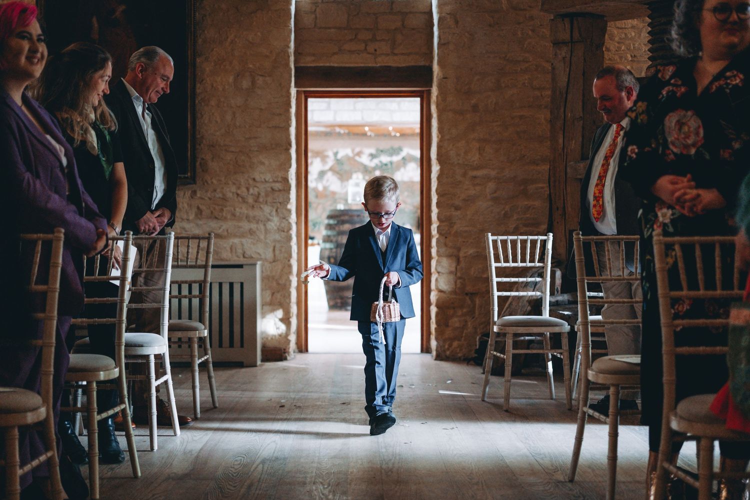 Flower boy strides through a rustic brick-walled venue with rows of chairs lining both sides, backlit by a bright doorway.