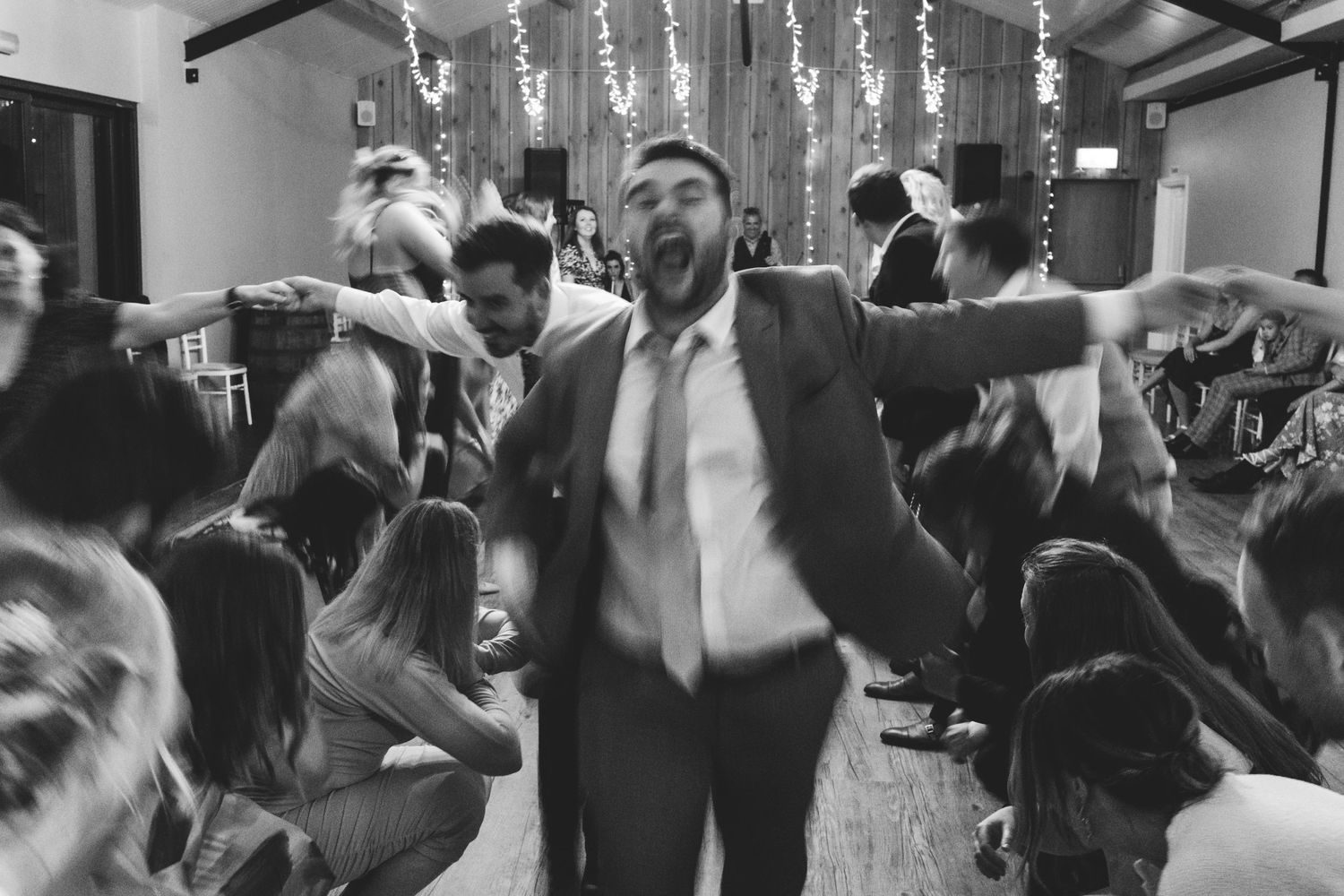 Black and white photo of people dancing enthusiastically at a wedding reception with hanging lights and festive decor.