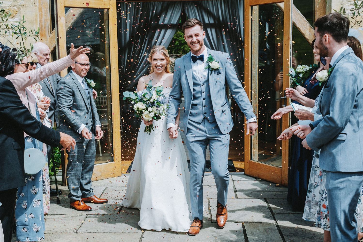 Wedding couple exits venue as guests shower them with confetti while wearing coordinated gray and blue attire.