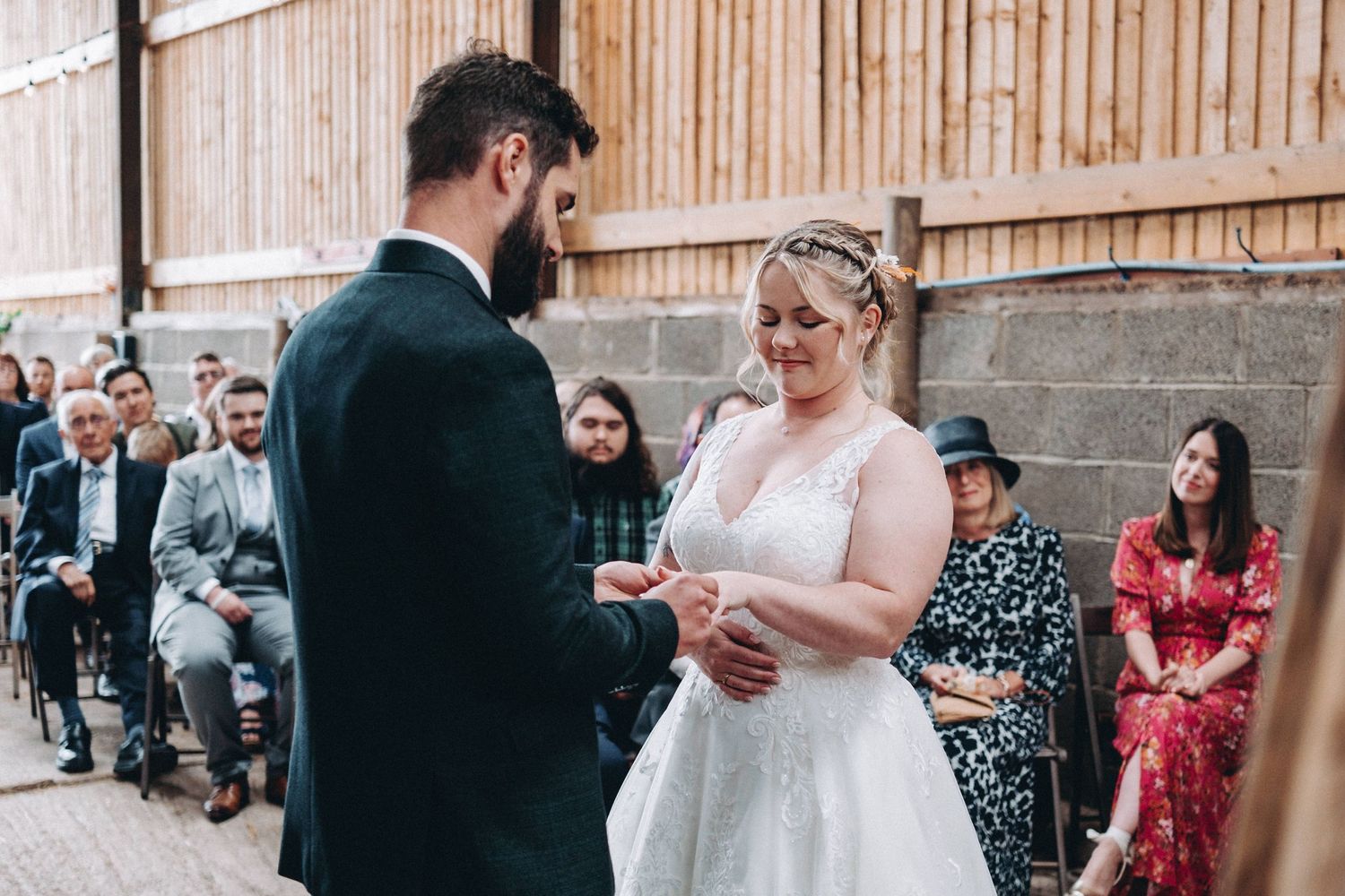 A couple exchanges rings during an intimate wedding ceremony in a rustic wooden venue with guests looking on.