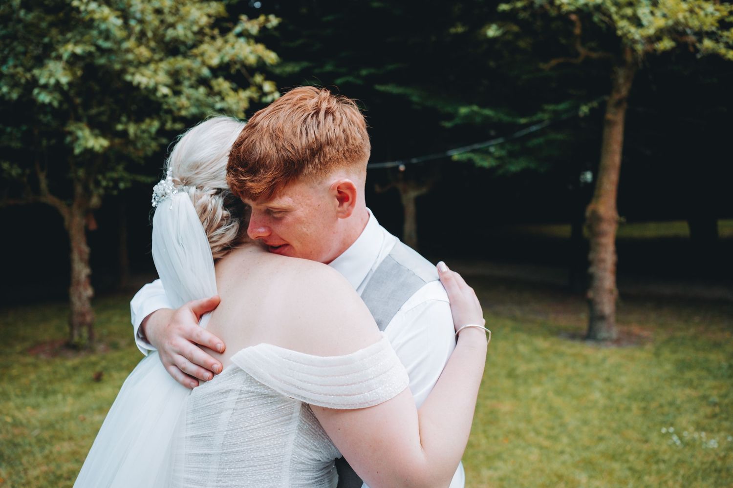 A tender embrace between bride and brother in wedding attire sharing an intimate moment outdoors in a wooded setting.
