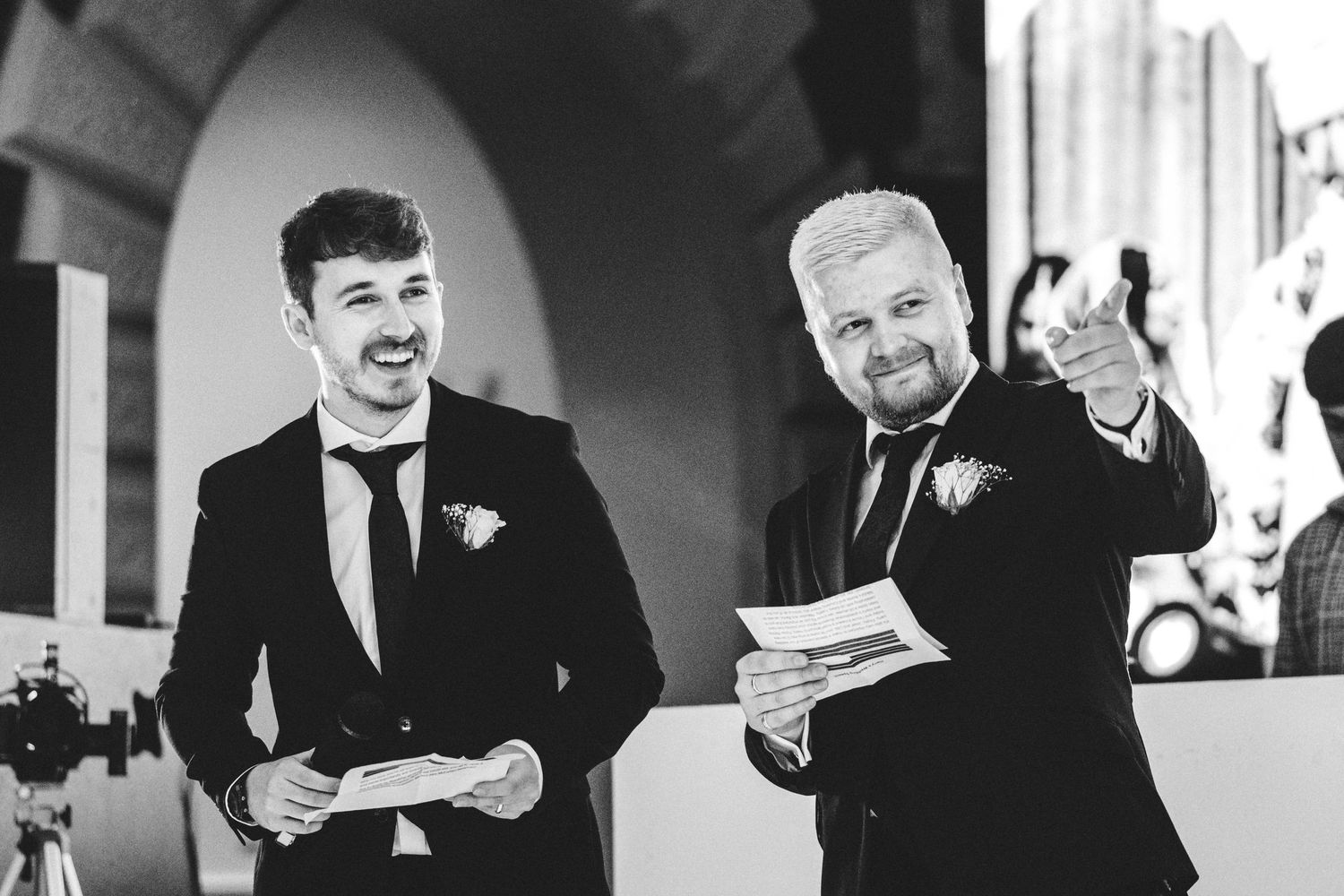 Wedding guests in formal attire share laughter while reading papers at an event.
