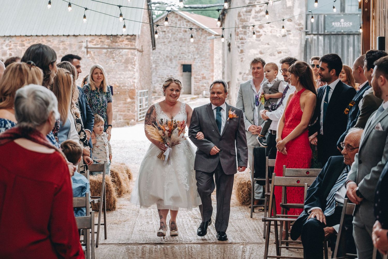 Guests look on as a couple walks down the aisle during a bright indoor ceremony with string lights and brick walls.