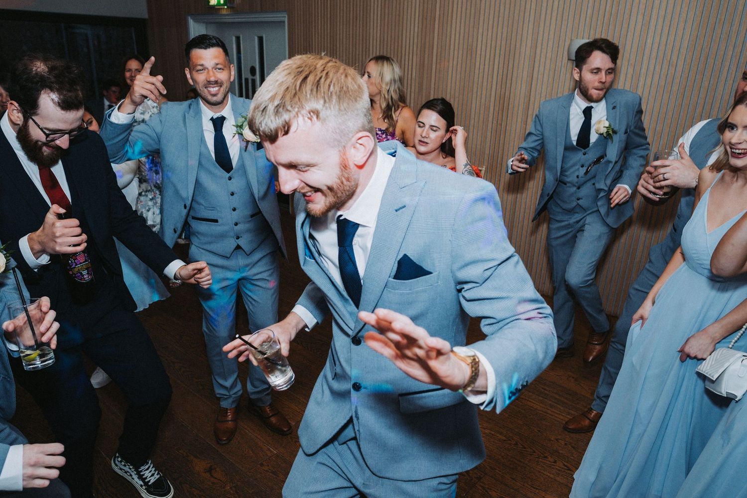 Wedding guests in matching light blue suits dance energetically at a reception celebration.