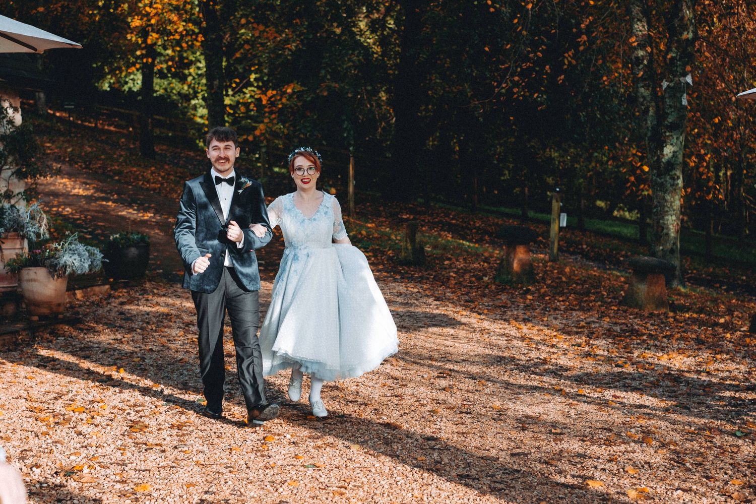A couple walks together through autumn leaves on a garden path in wedding attire.