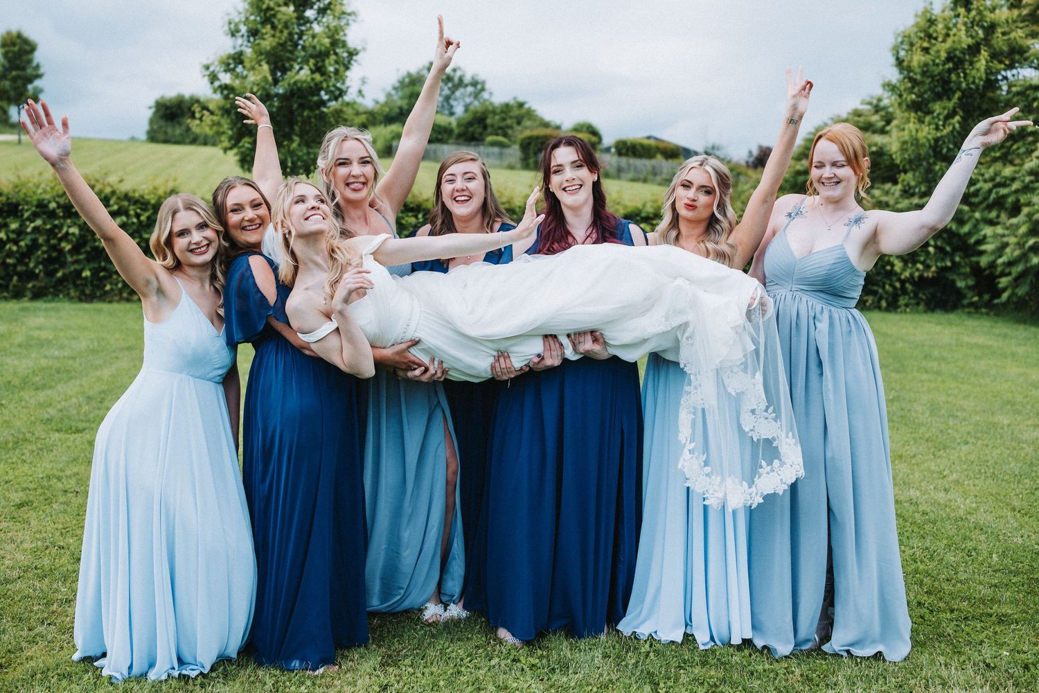 Bridesmaids in varying shades of blue dresses lift and support someone in a white gown on a sunny lawn.