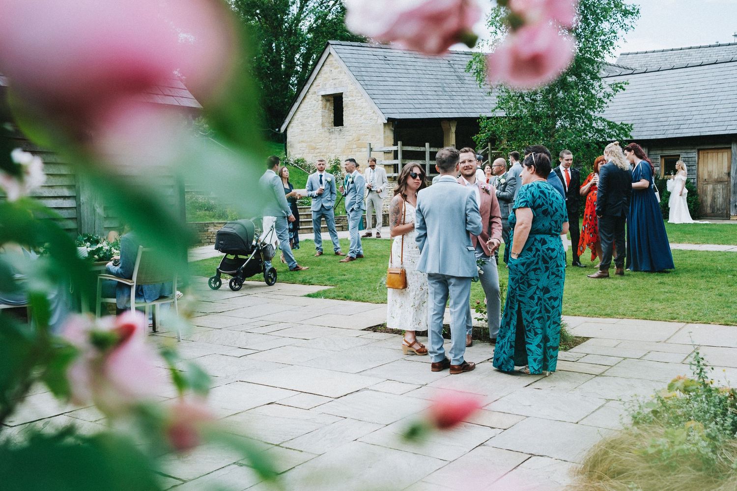 Wedding guests mingle on a stone patio outside a rustic venue with pink flowers in the foreground.