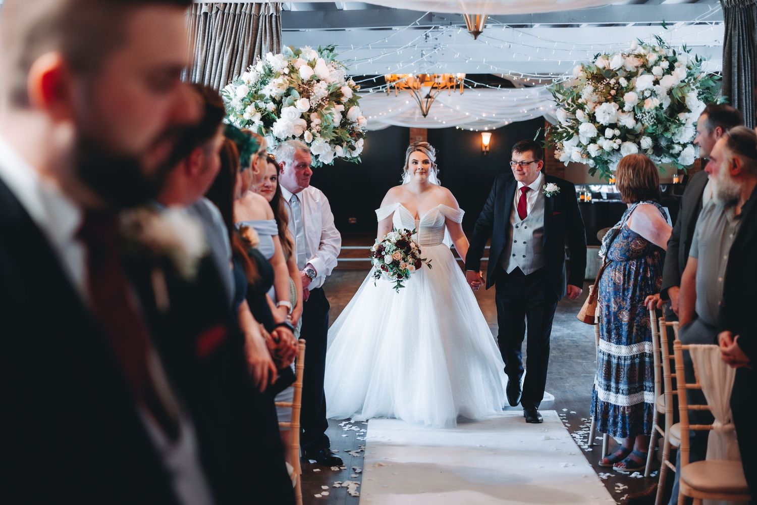 Wedding ceremony setup with floral arch and white aisle runner as guests gather at an elegant indoor venue.