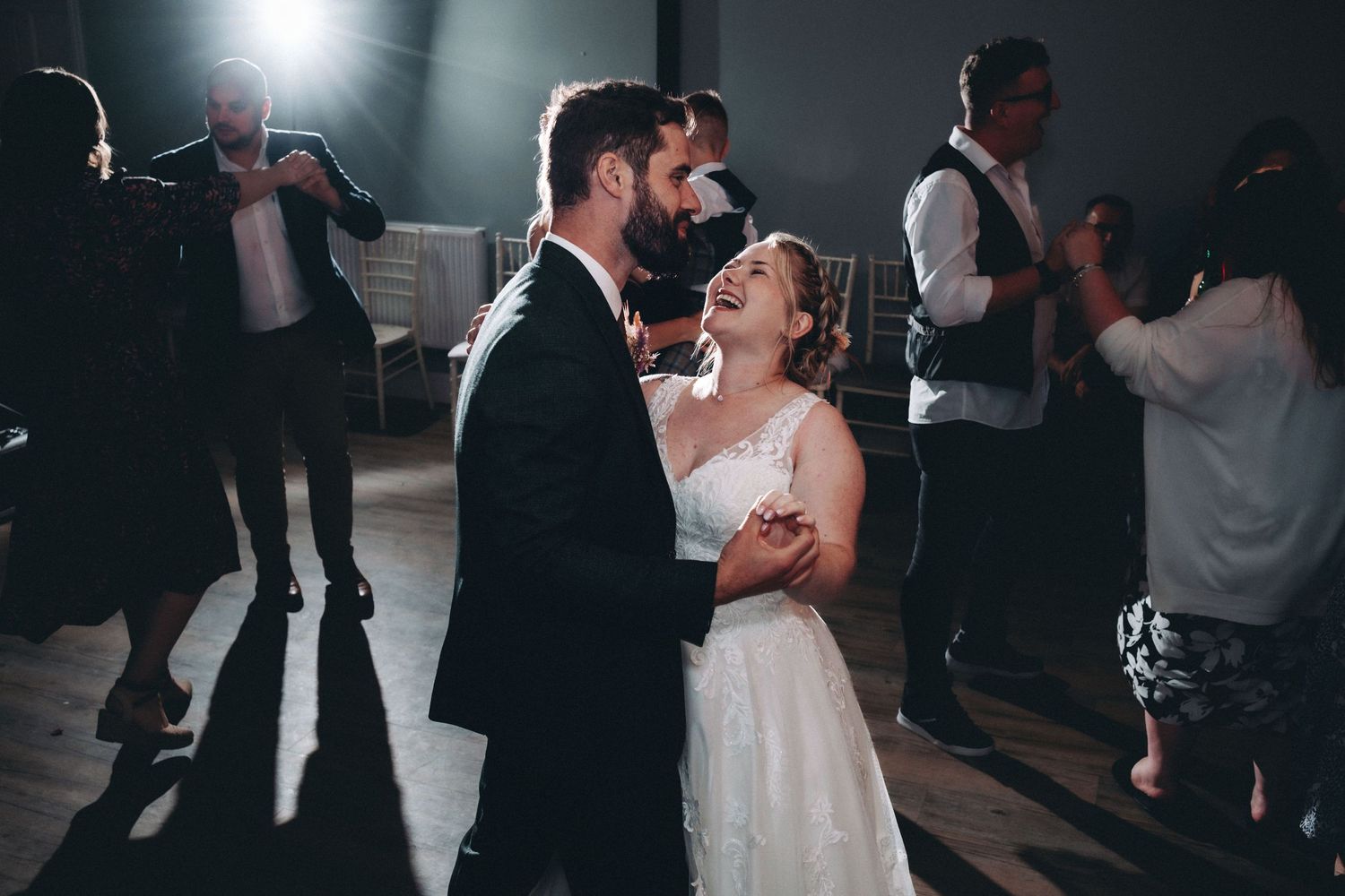 A couple shares a joyful moment while dancing at their wedding reception with a bright backlight and guests in the background.