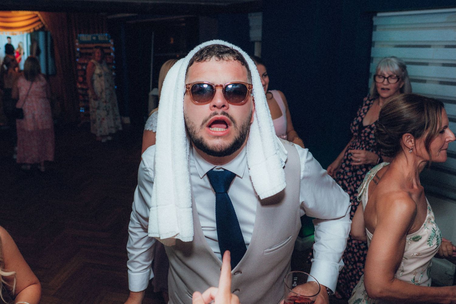 Party guest wearing sunglasses and a white towel on head dances energetically at indoor celebration.