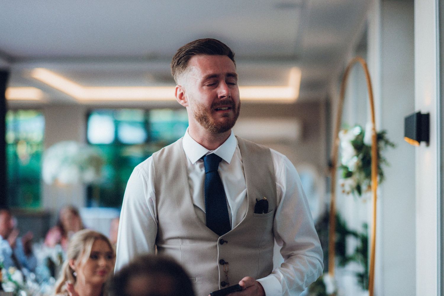 A person in a light colored vest and blue tie speaking during a wedding reception.